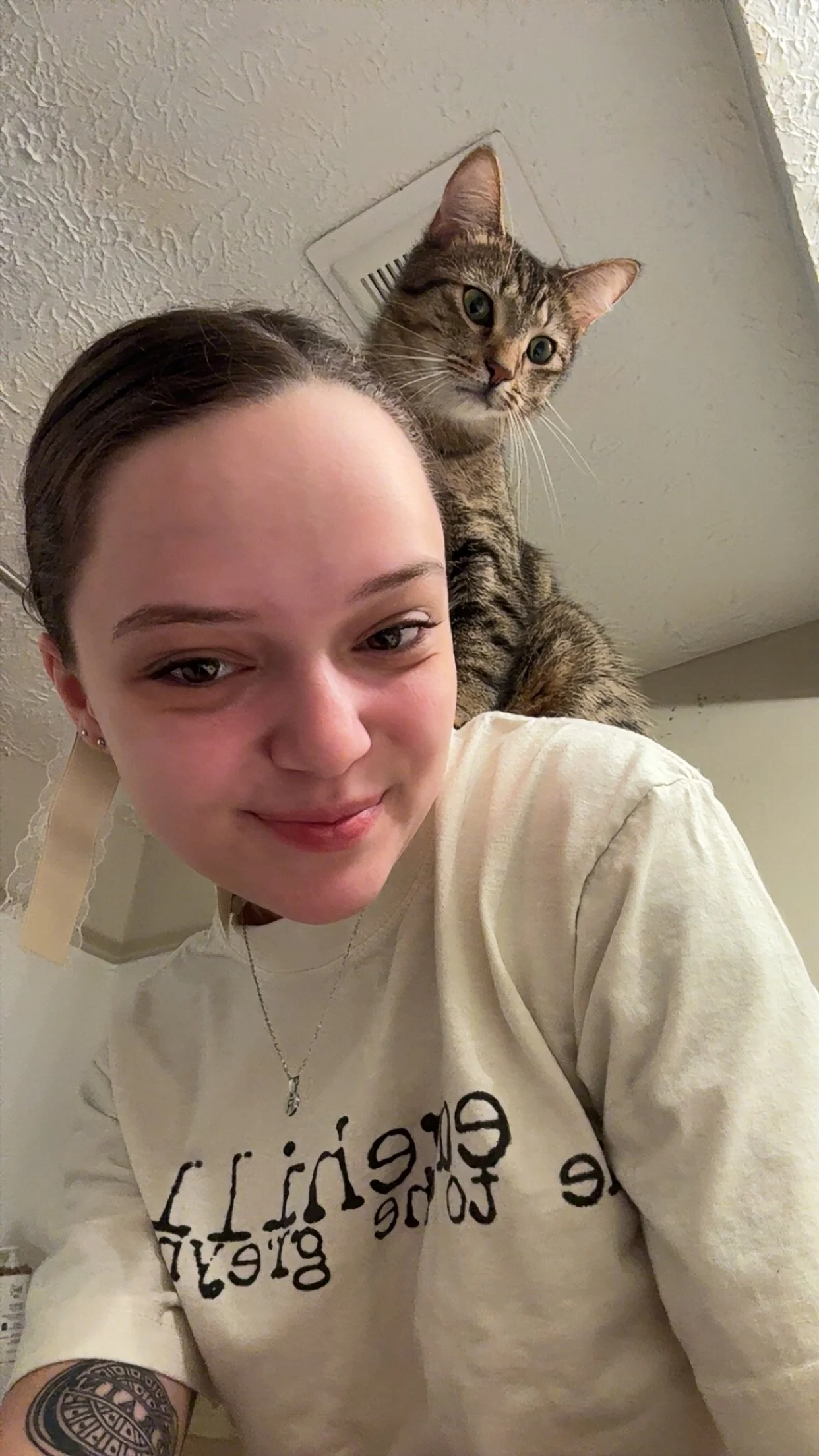 A young woman with short hair, earrings, and a necklace smiling at the camera. She is wearing a cream-colored sweatshirt with black lettering. Behind her, a tabby cat with green eyes is perched on her shoulder, looking into the camera. The background features a textured white ceiling with a vent.