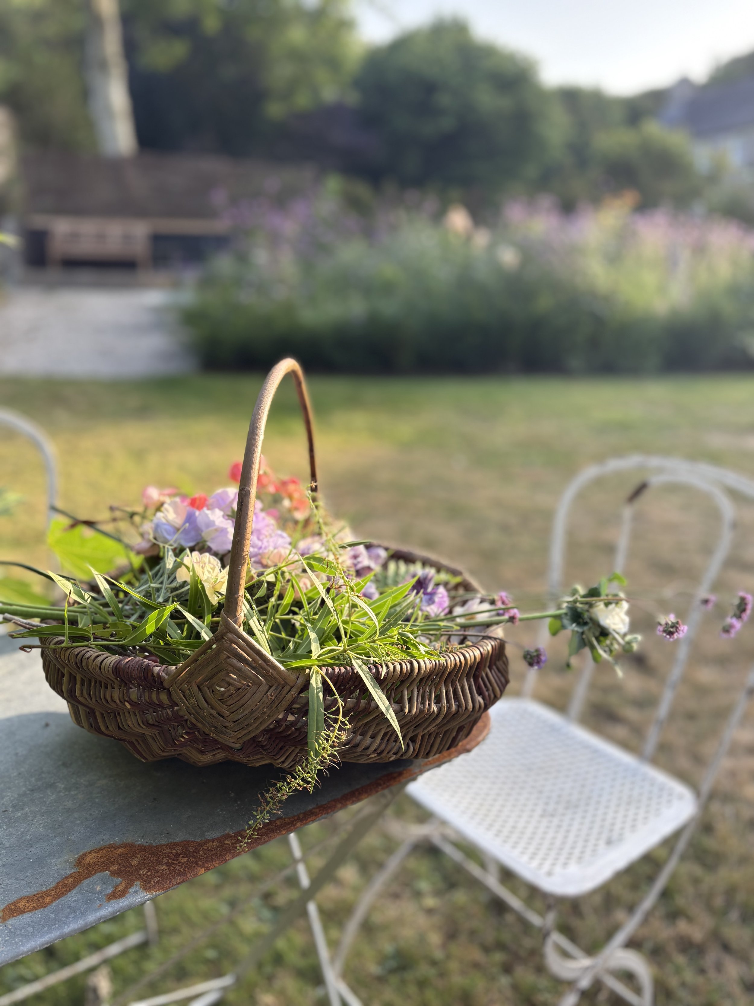 A wicker basket filled with colorful flowers resting on an outdoor table, with a white metal chair and garden background.