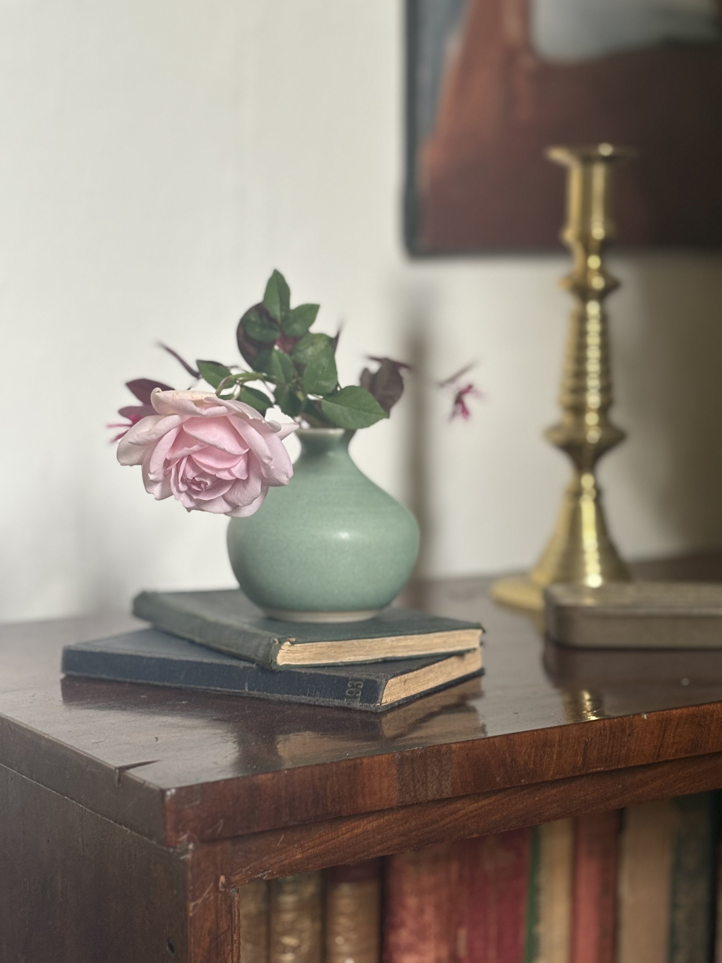 A pink rose in a small green ceramic vase placed on two old hardcover books on a wooden surface. In the background, a gold candlestick and a blurred framed picture are visible.