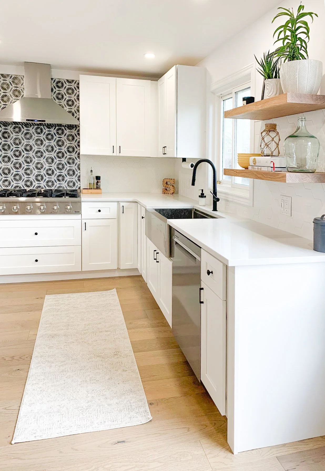 Modern kitchen with white cabinets, patterned backsplash, stainless steel stove and range hood, farmhouse sink, open wooden shelves with plants and decor, and light wood flooring.