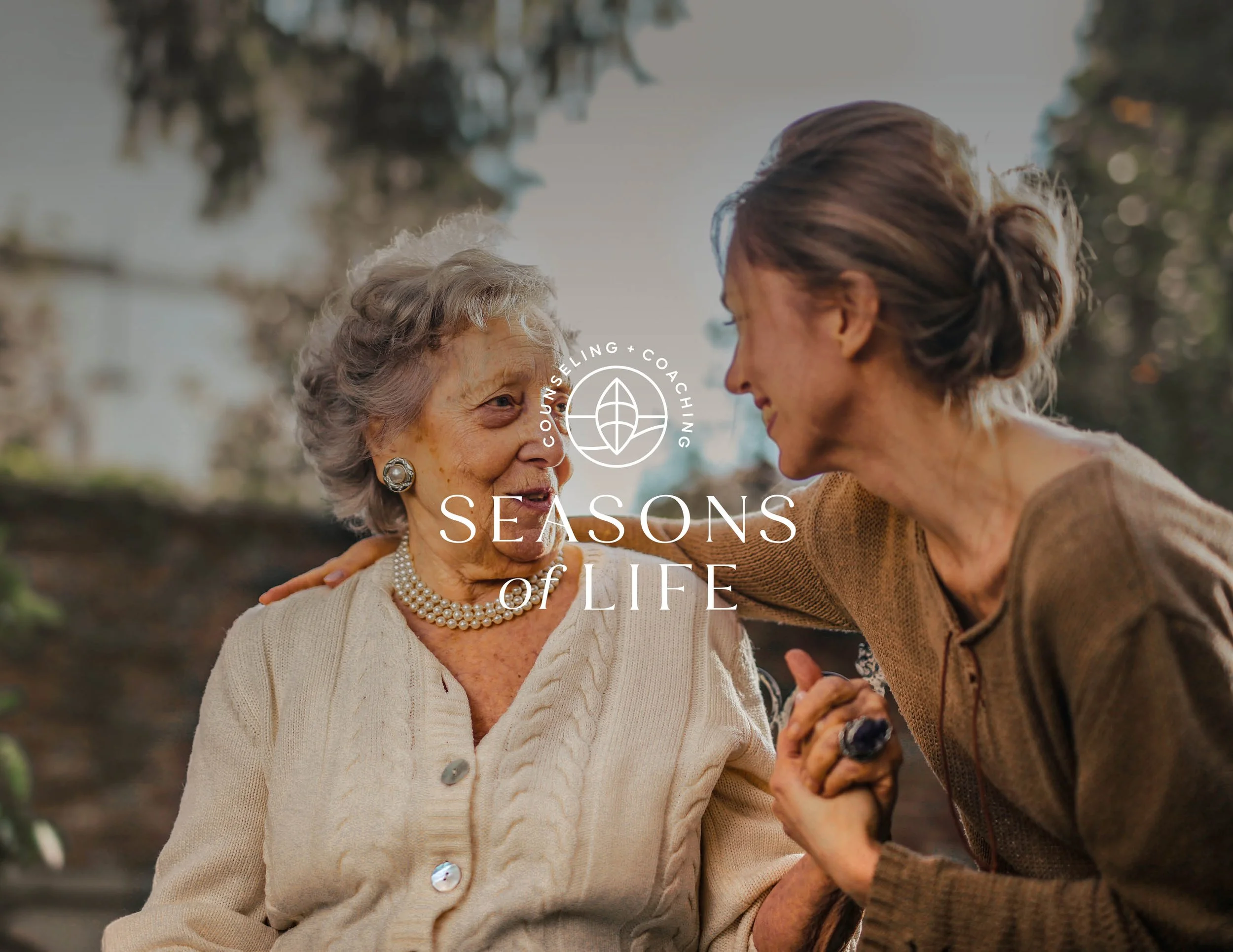Two women, an elderly woman and a middle-aged woman, smiling and holding hands outdoors, with trees in the background.