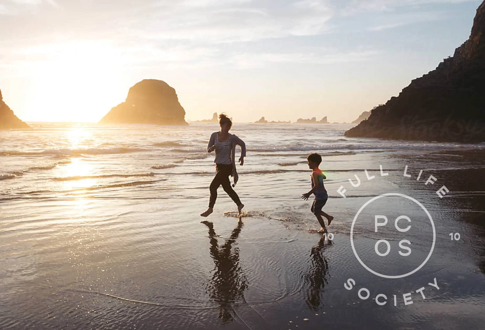 A person and a child running on a beach at sunset with rocks in the background.