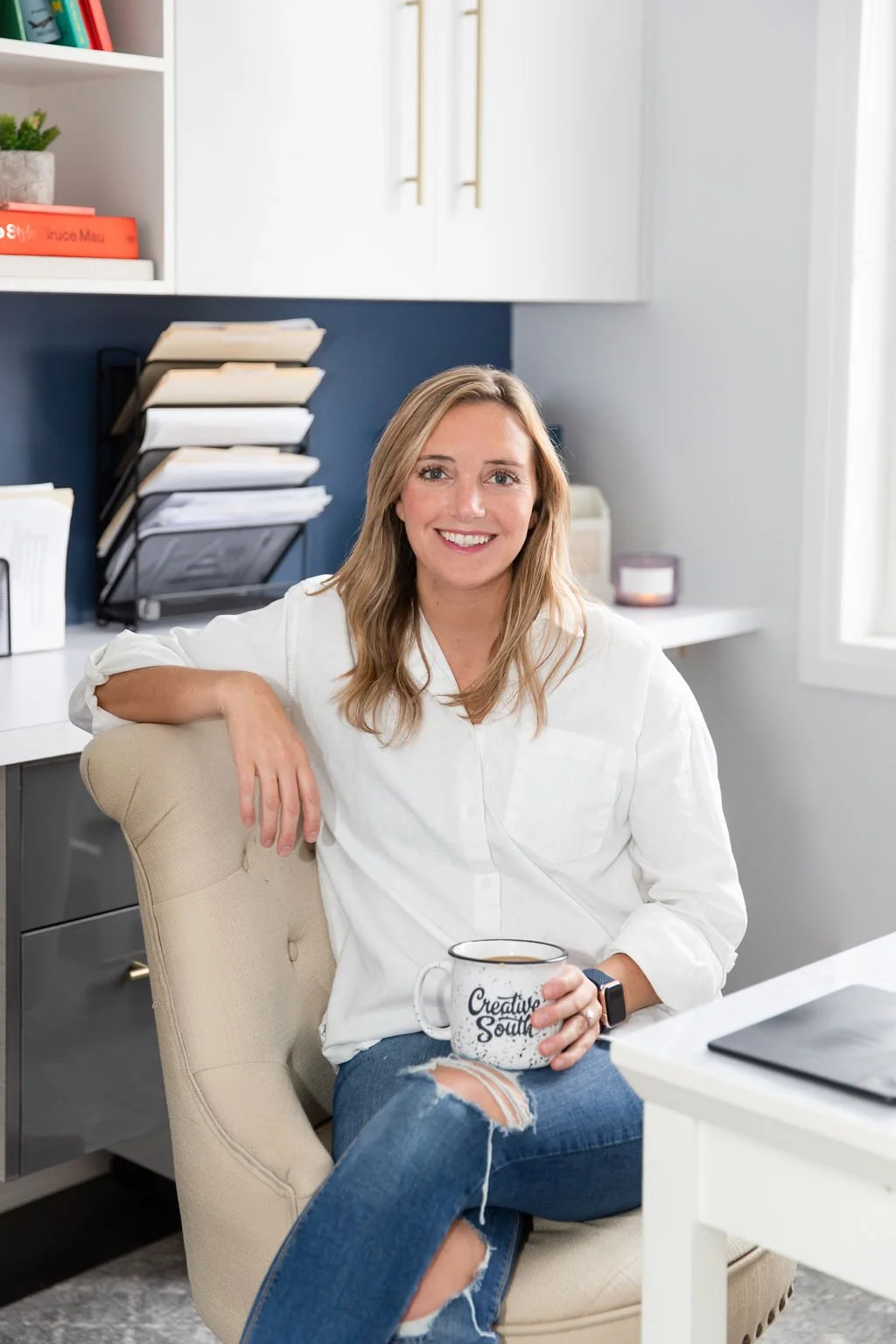 A woman sitting in a beige chair at a white desk in an office, holding a coffee mug, smiling, with desk and storage shelves in the background.