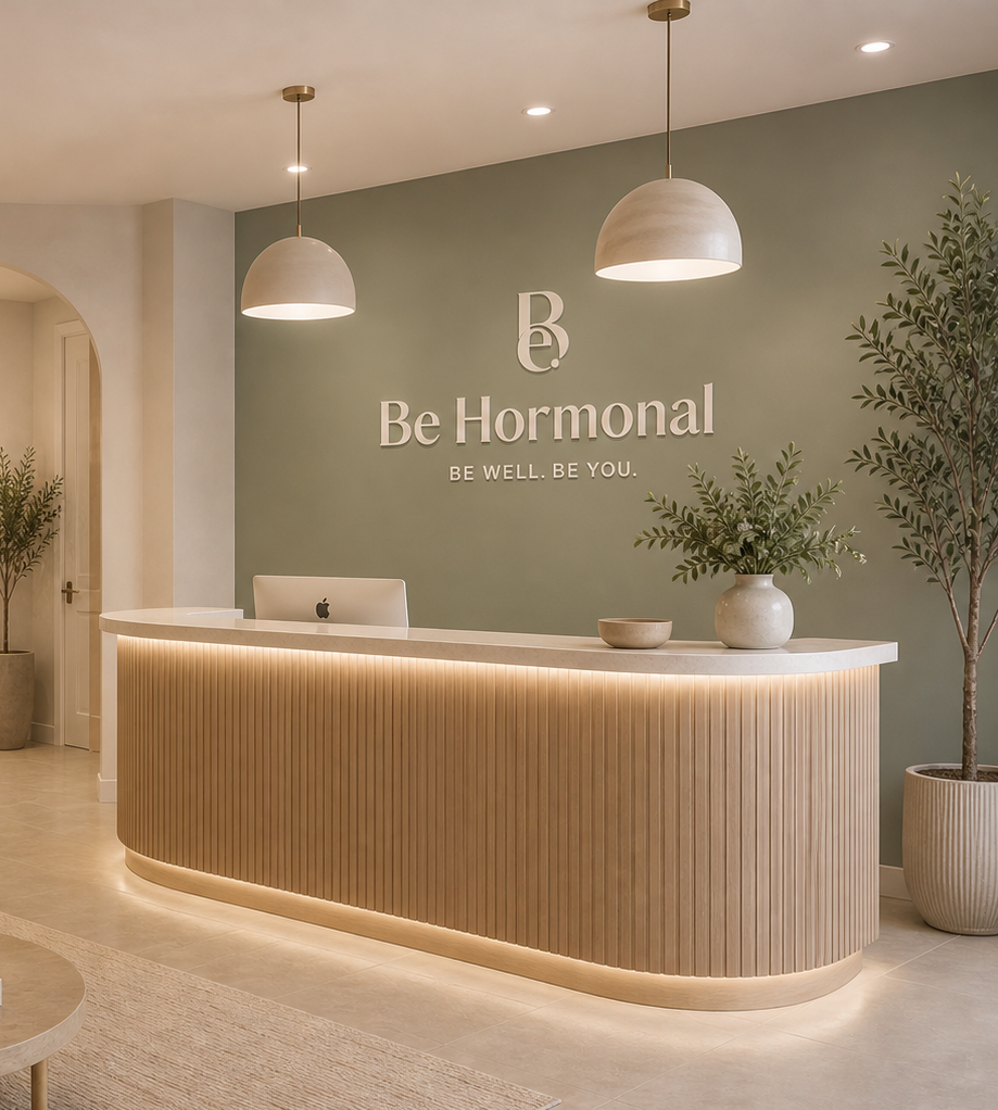 Modern reception area with a curved wooden desk, a desktop computer, and decorative plants, with a mint green wall featuring the logo and name 'Be Hormonal' and the tagline 'Be Well. Be You.' Two hanging pendant lights illuminate the space.