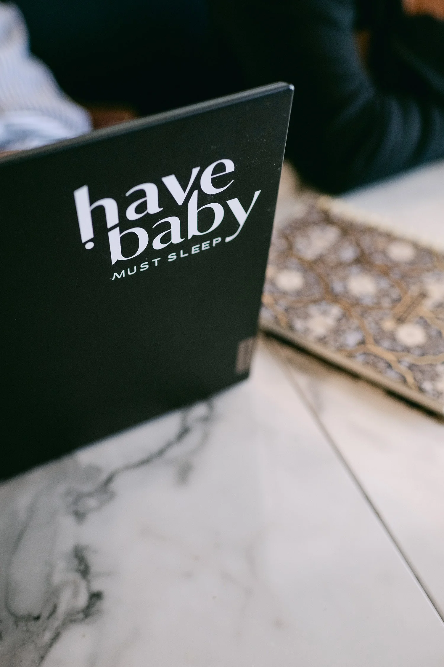 Close-up of a black book titled '!have baby MUST SLEEP' on a marble table, with someone sitting in the background.