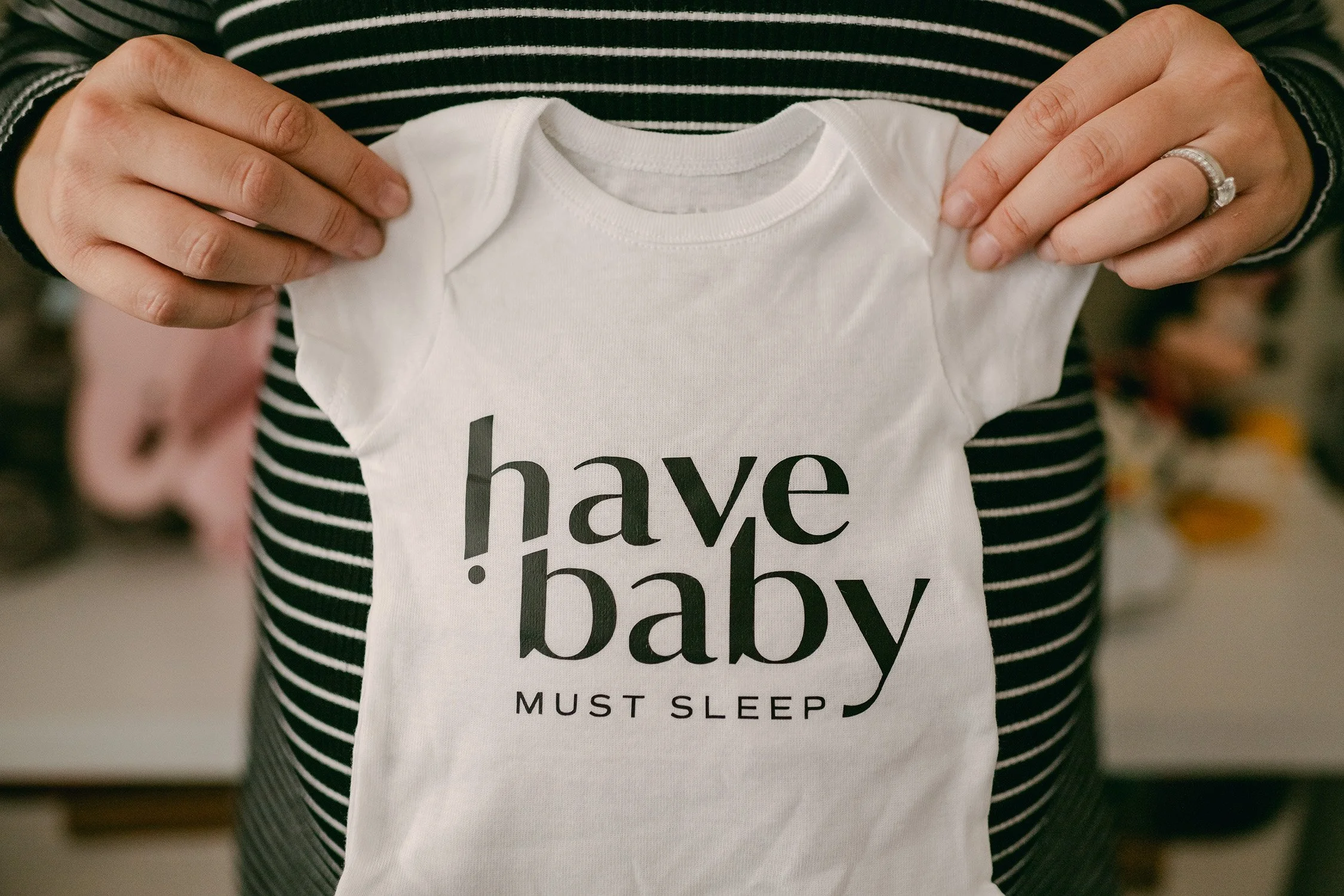 A person holding a white baby onesie with the text "have! baby" and above it, "MUST SLEEP" written in black, with a background of a person in a black and white striped shirt.