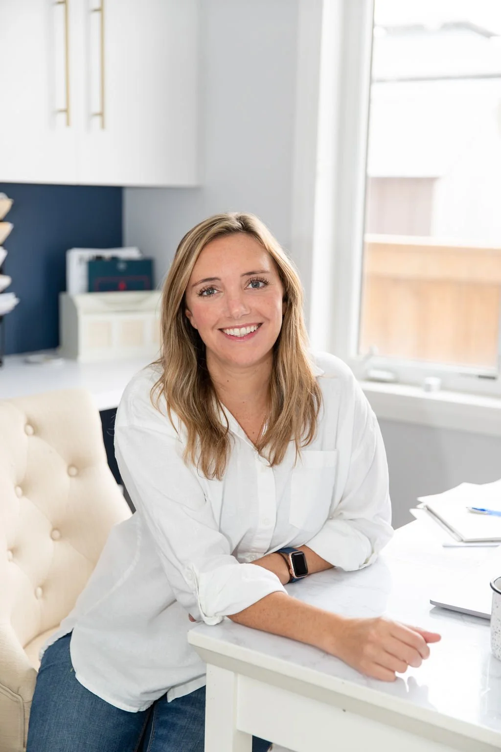 A woman with long light brown hair smiles while sitting at a desk in a bright office.
