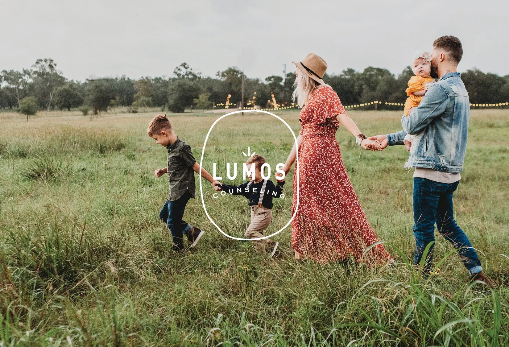 A family walking hand in hand through a grassy field. The mother wears a red floral dress and hat, the father holds a baby, and two children walk ahead. Lumos Counseling logo is overlaid.