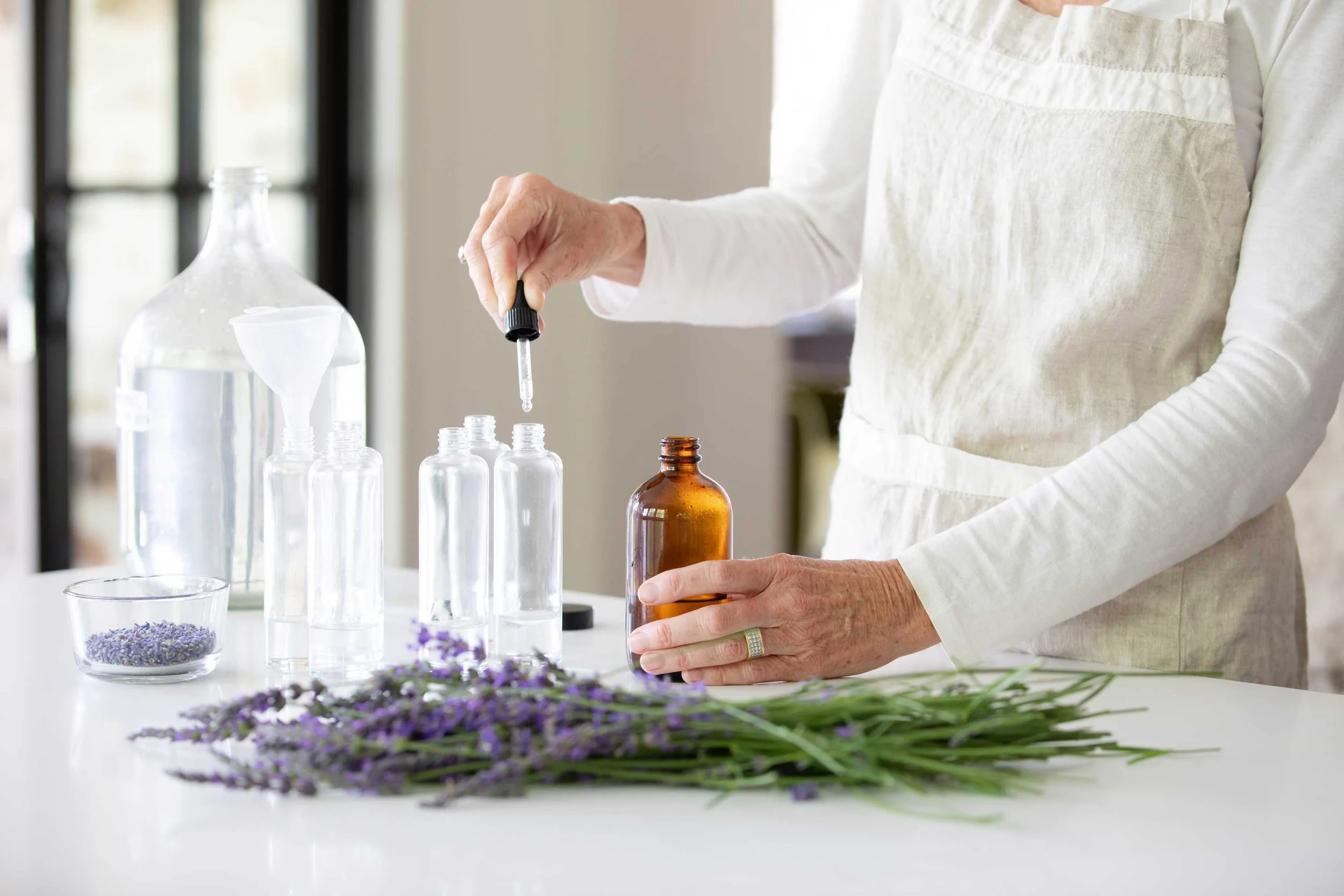 Person preparing essential oils with a dropper, clear and amber bottles, lavender sprigs on a white surface.