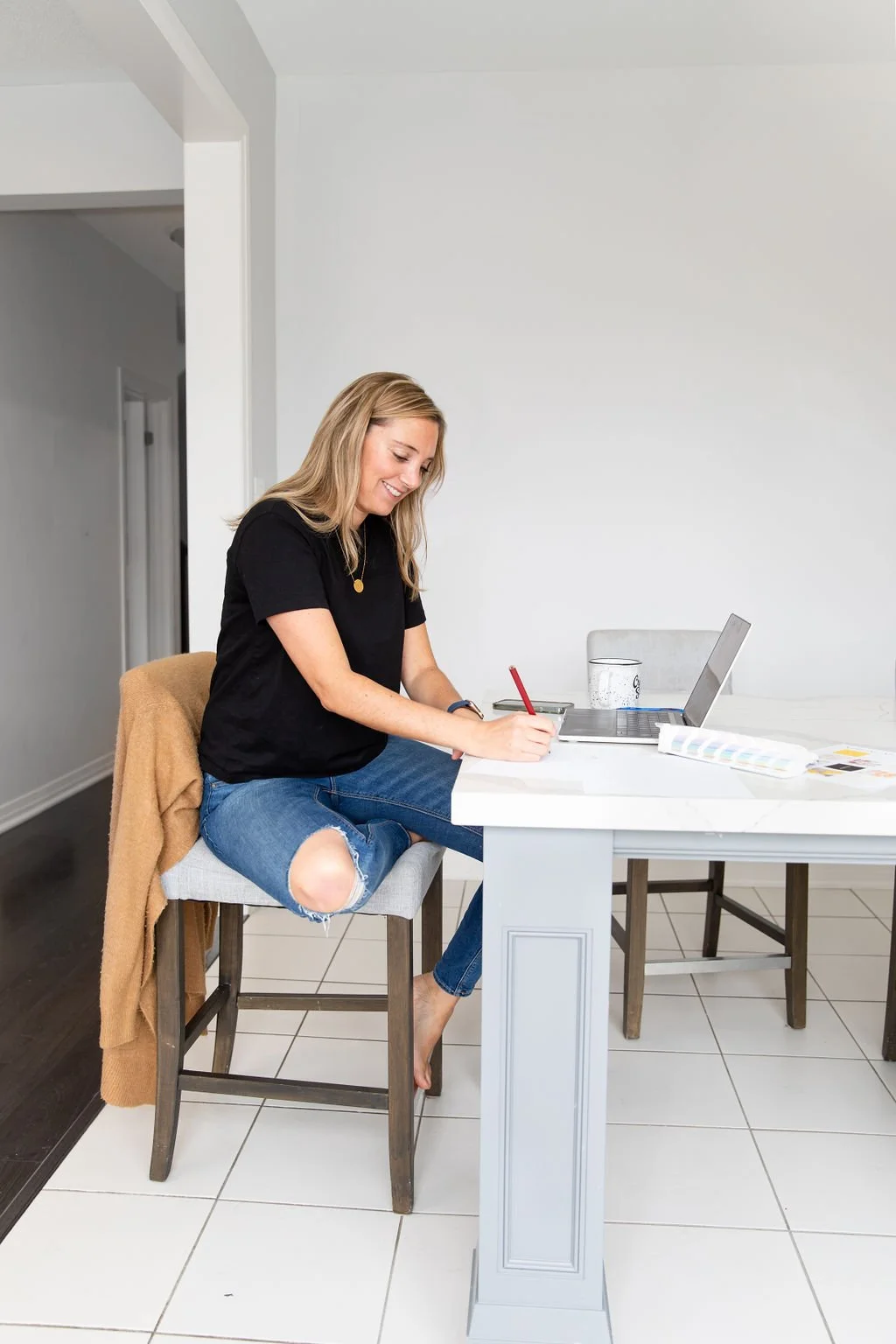 A woman with blonde hair is sitting on a barstool at a kitchen island, writing with a red pen. She is smiling, wearing a black t-shirt and ripped jeans. There is a laptop, a glass of water, and some papers on the counter.