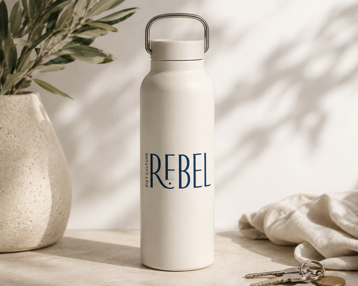 A white water bottle with the word 'REBEL' printed on it in blue, placed on a wooden surface next to a beige fabric, keys, and a potted plant with green and white leaves.