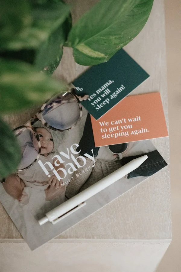 A baby sleep magazine, a pair of glasses, a white marker, and two cards with messages about sleep are placed on a surface near a green houseplant.