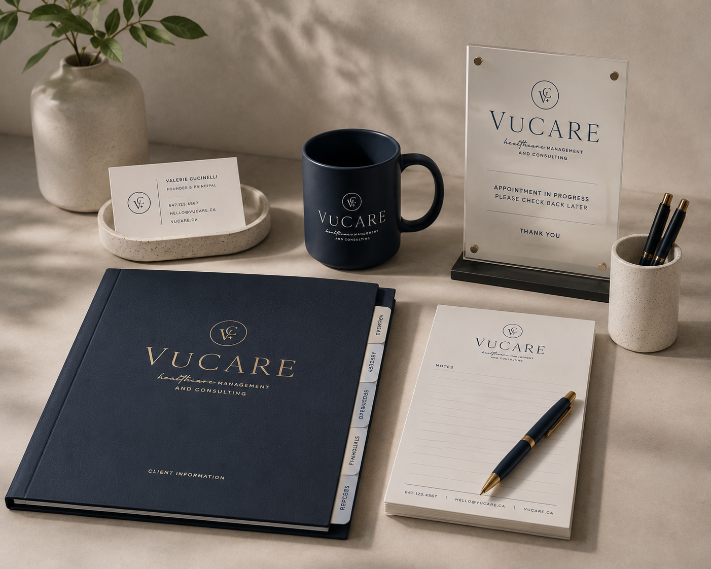 Office desk with VUCARE branding materials including a navy notebook, a white notepad with a pen, business cards, a navy mug, a clear sign, a cup with pens, and a vase with a plant.