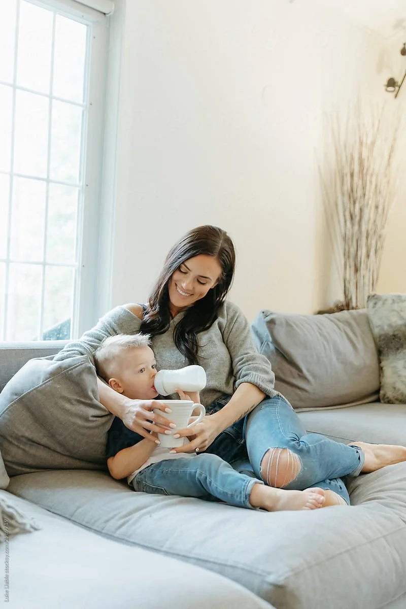 A woman sitting on a couch with a young child drinking from a bottle, both smiling, in a bright living room with a window and neutral decor.