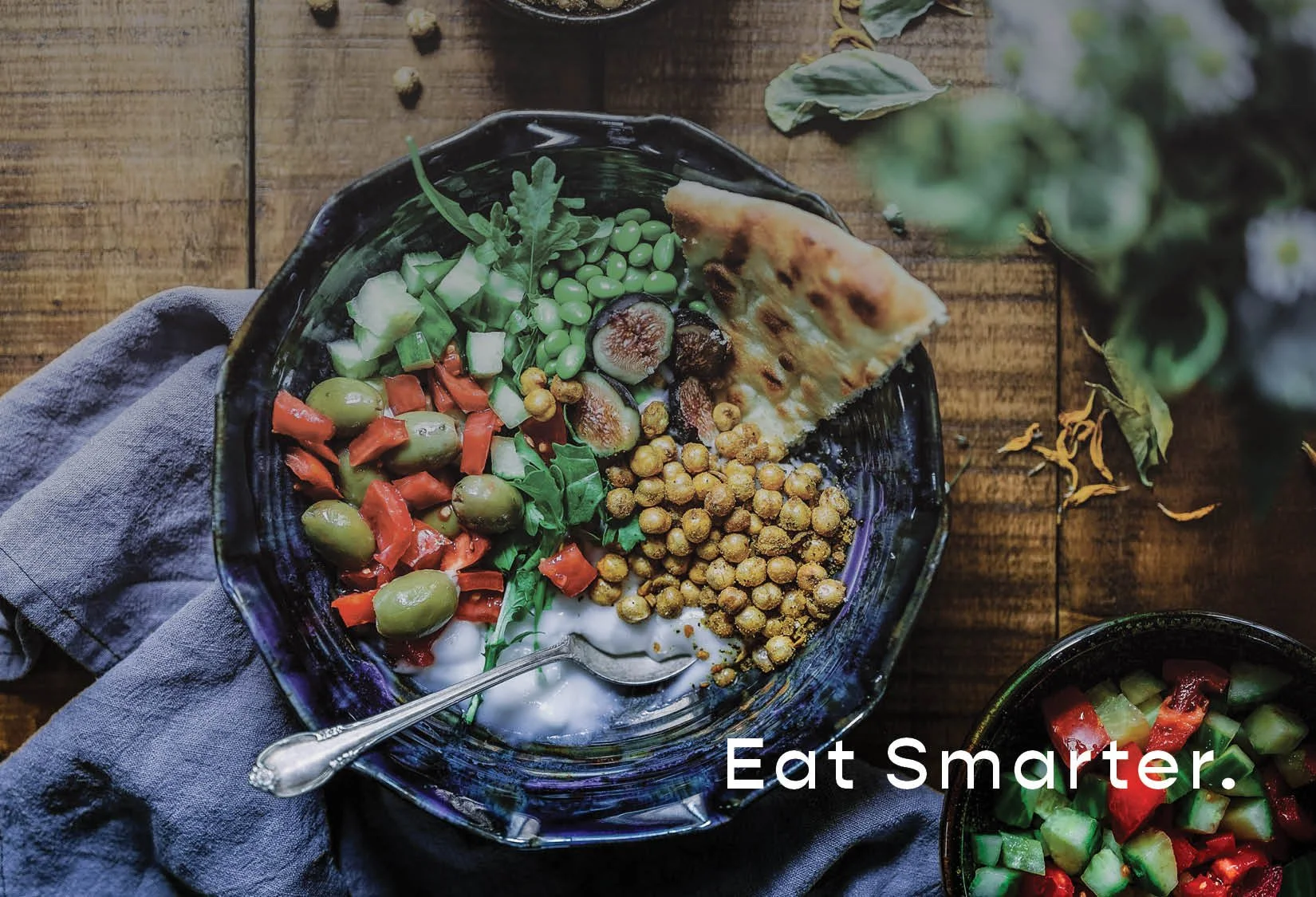 Bowl of mixed salad with chickpeas, cucumbers, olives, figs, edamame, arugula, red peppers, and a piece of flatbread, placed on a wooden table with a smaller bowl of cucumber and red pepper salad, accompanied by the text 'Eat Smarter.'