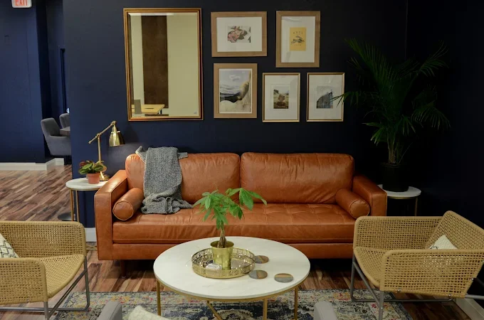 Living room with a brown leather sofa, framed artwork on dark wall, side tables with plants, and rattan chairs.