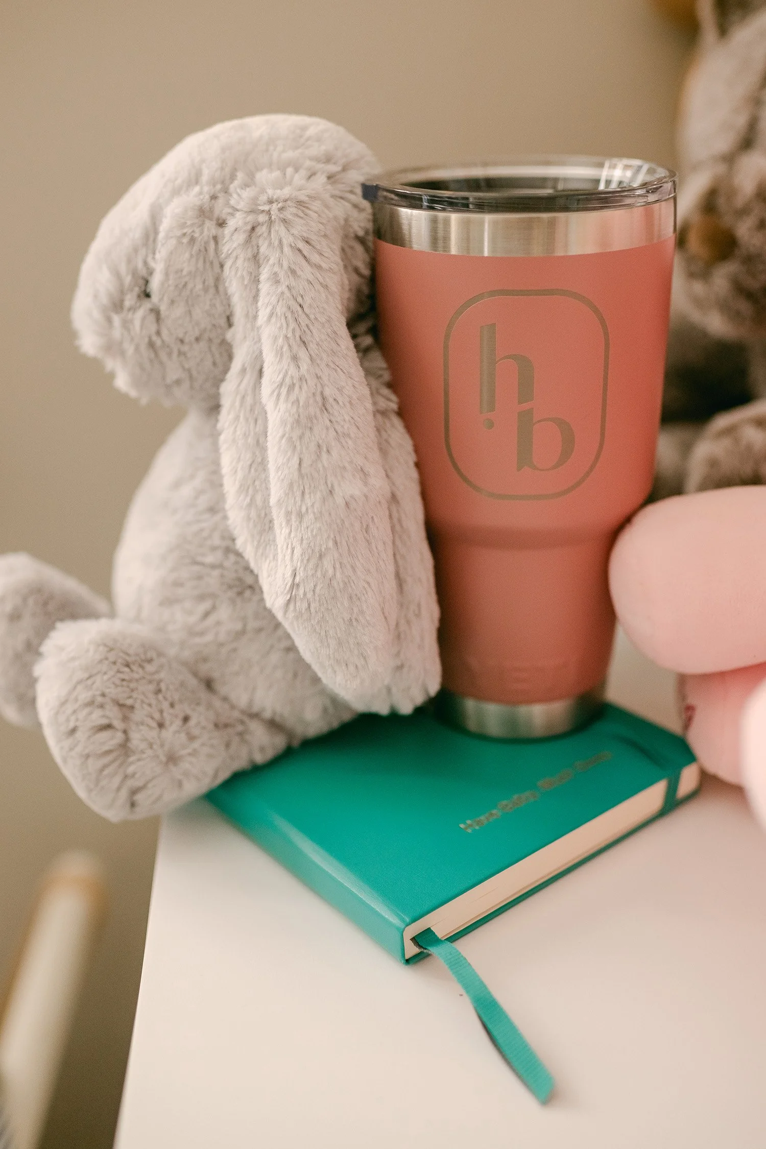 A plush gray stuffed bunny, a pink tumbler with the logo 'h.b', and a teal book with a ribbon bookmark on a white surface.