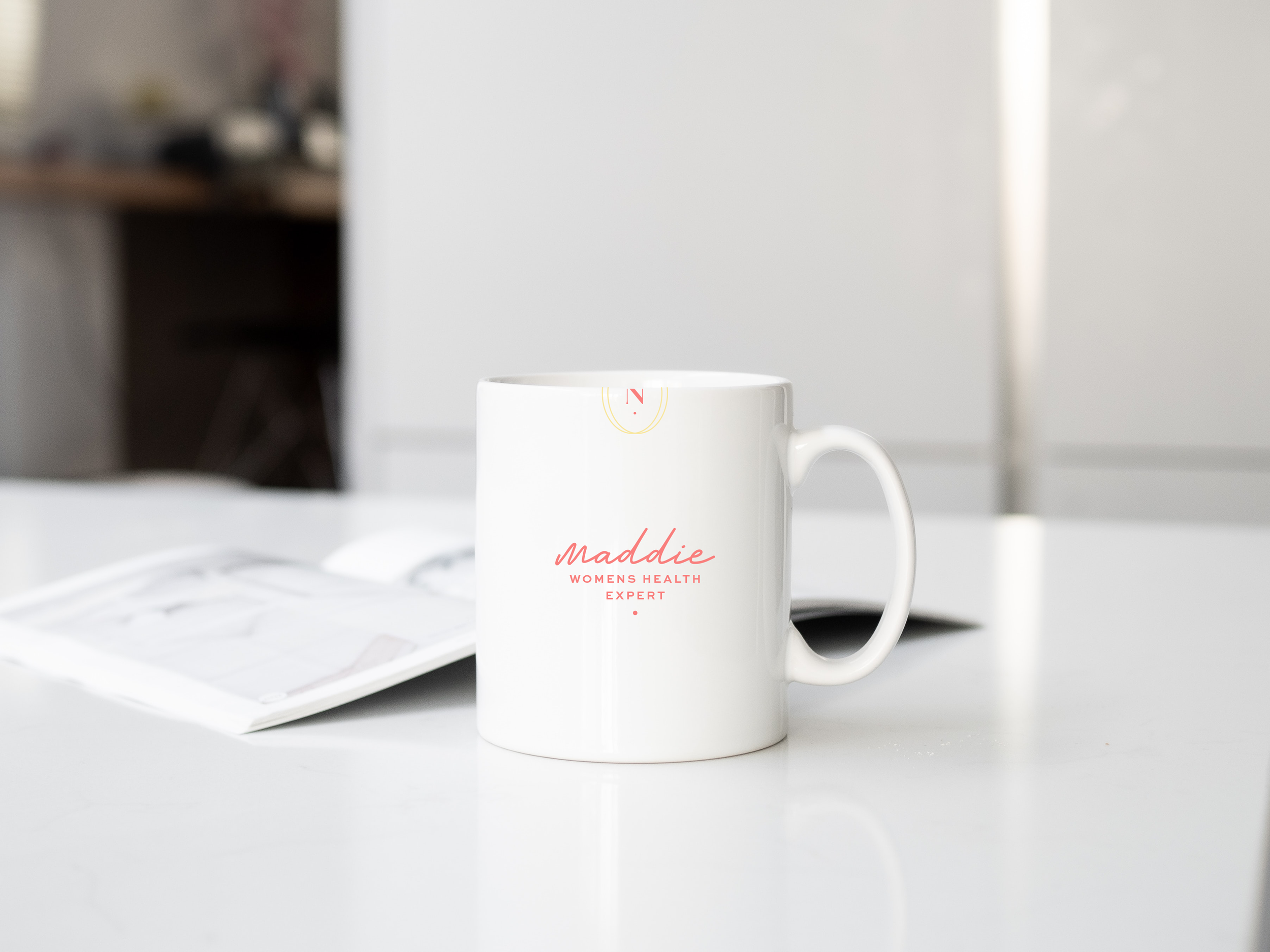 White coffee mug on table with text "Maddie Women's Health Expert" in pink and red lettering, open magazine in background.