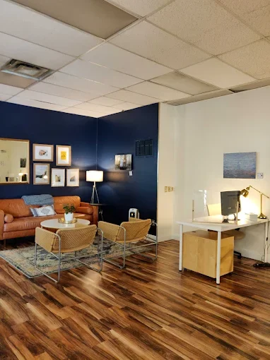 A cozy office lounge with a blue accent wall, framed artwork, a tall floor lamp, a brown sofa, and two tan chairs on a patterned rug. Adjacent is a white desk with a swivel chair, computer, lamp, and wall art.