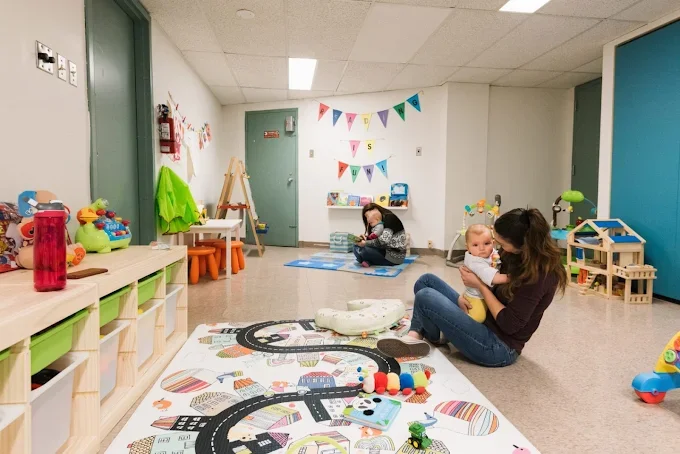 A cozy indoor playroom with children and adults. The room has colorful toys, a play mat with roads, and decorations on the wall, including a banner and artwork. One adult is holding a baby, and another is kneeling on the floor."
