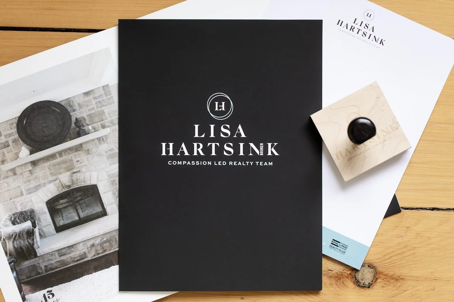 Business brochure and promotional materials for Lisa Hartsink, a realtor, on a wooden table. The main brochure is black with white text and a logo. There are additional papers, including a white sheet and a photo of a living room interior.