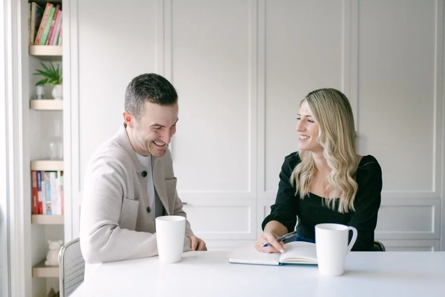A man and woman sitting at a white table, smiling and having a conversation in a bright room, with books and a plant in the background.