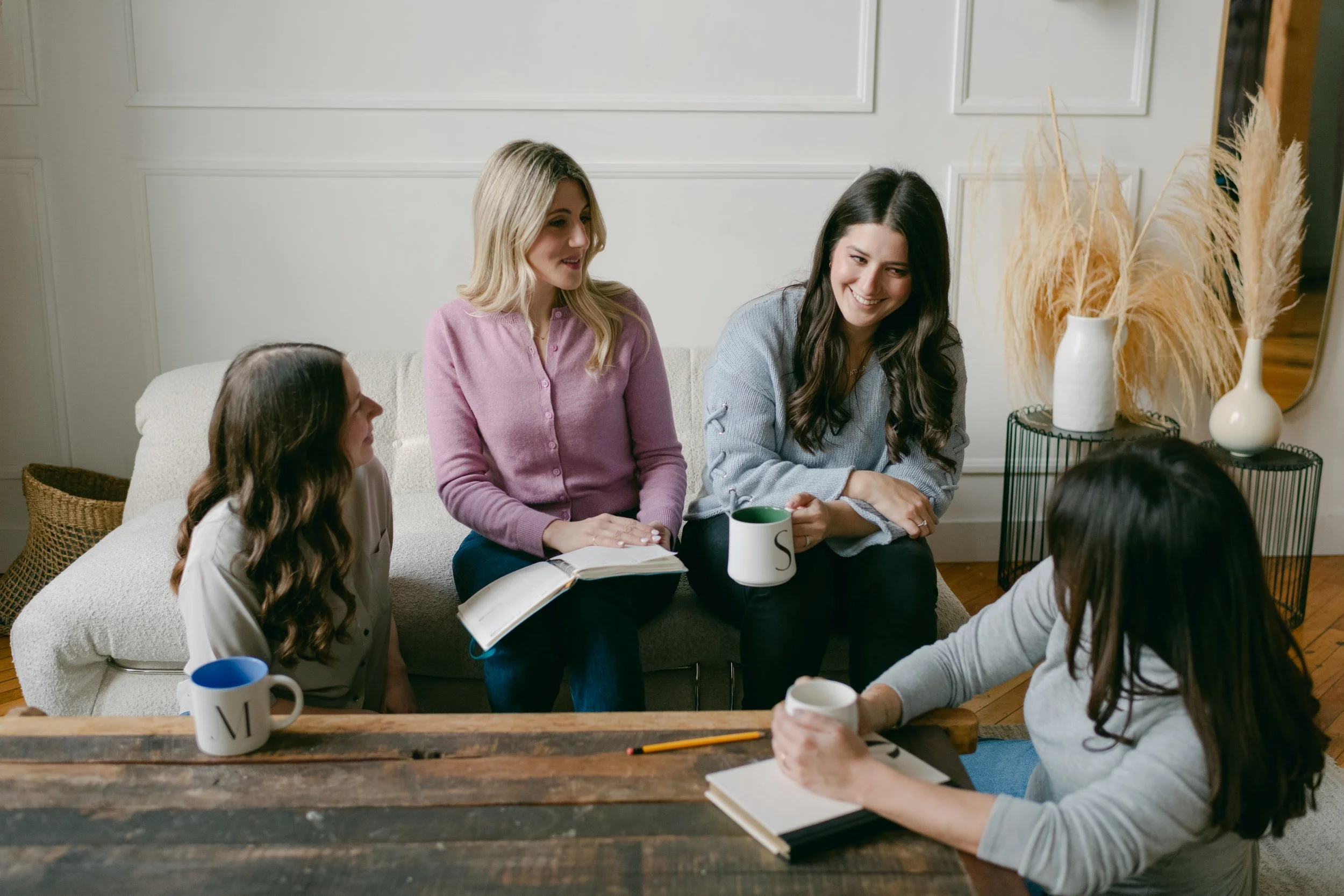 Group of women sitting together in a living room, engaging in conversation with coffee mugs and notebooks on the table.