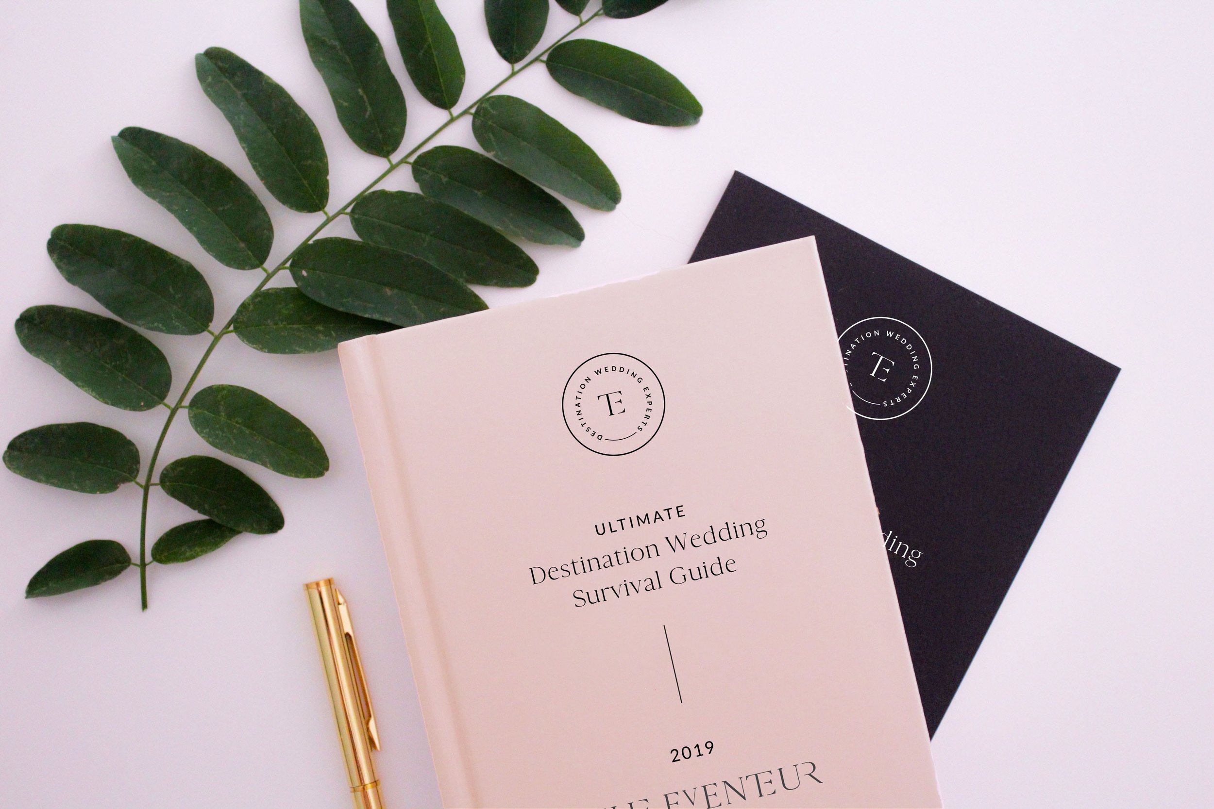 A sprig of green leaves, a gold pen, and two event guide booklets titled "Ultimate Destination Wedding Survival Guide" placed on a white surface. One booklet is white with black text, and the other is black with white text.