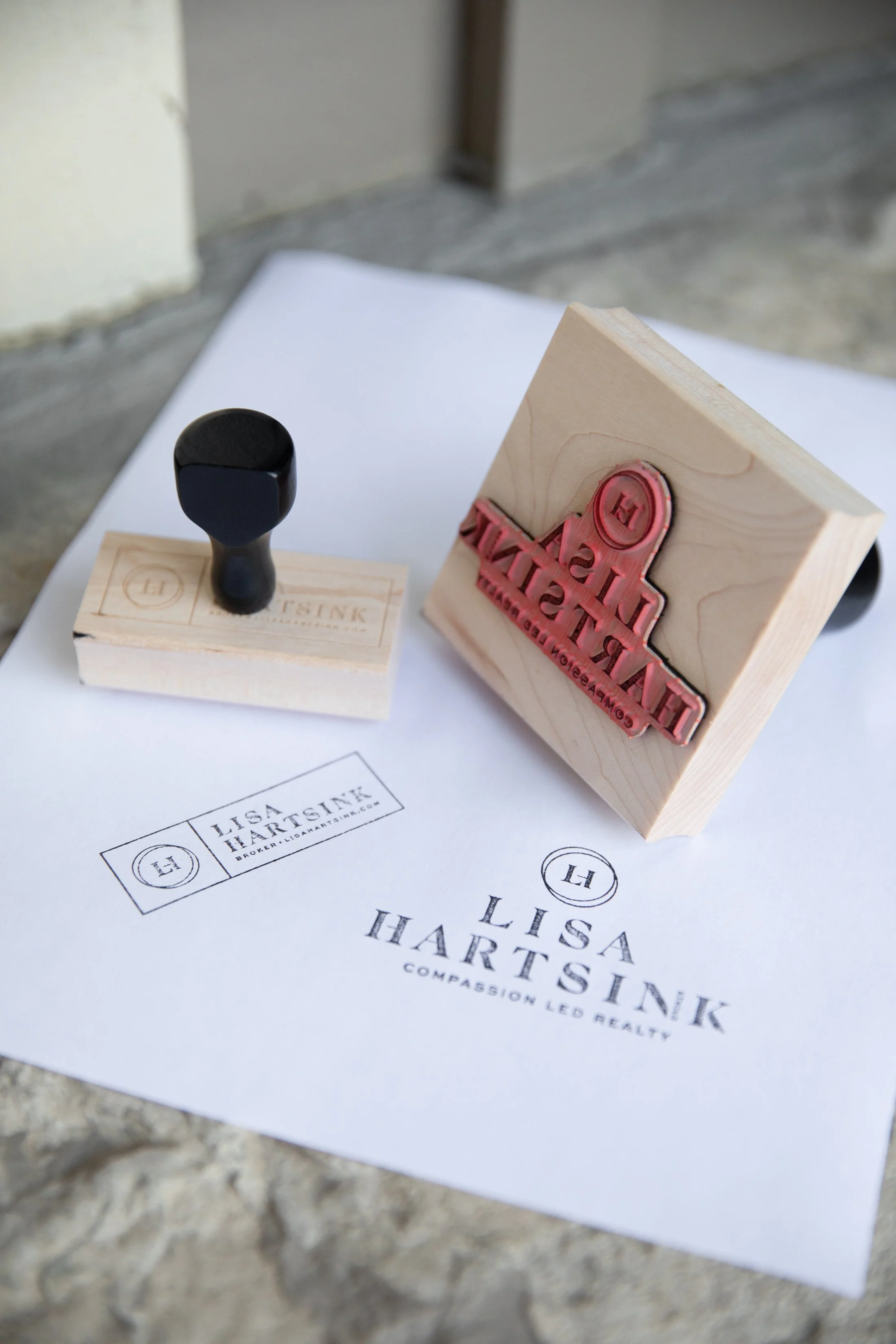 Two wooden rubber stamps with black handles on a paper displaying stamped text reading 'Lisa Hartsink' and 'Compassion Led Realty.'"