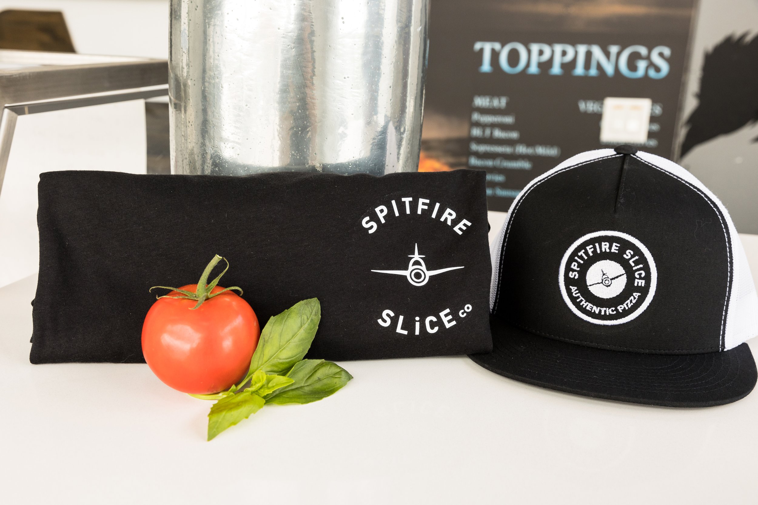 Black t-shirt and hat with "Spitfire Slice Co" logo next to a tomato and basil leaves on a white surface, with a toppings menu in the background.