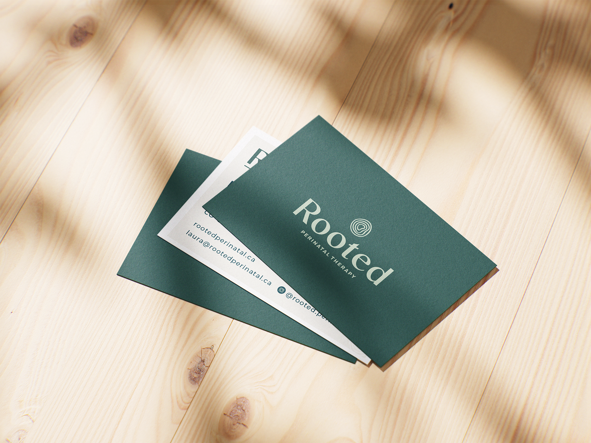 Business cards on a wooden surface, one card is green with white text and logo, with the word 'Rooted' visible, and the other is white with black text. The green card features contact details and social media handle, indicating a therapy or wellness business called Rooted.
