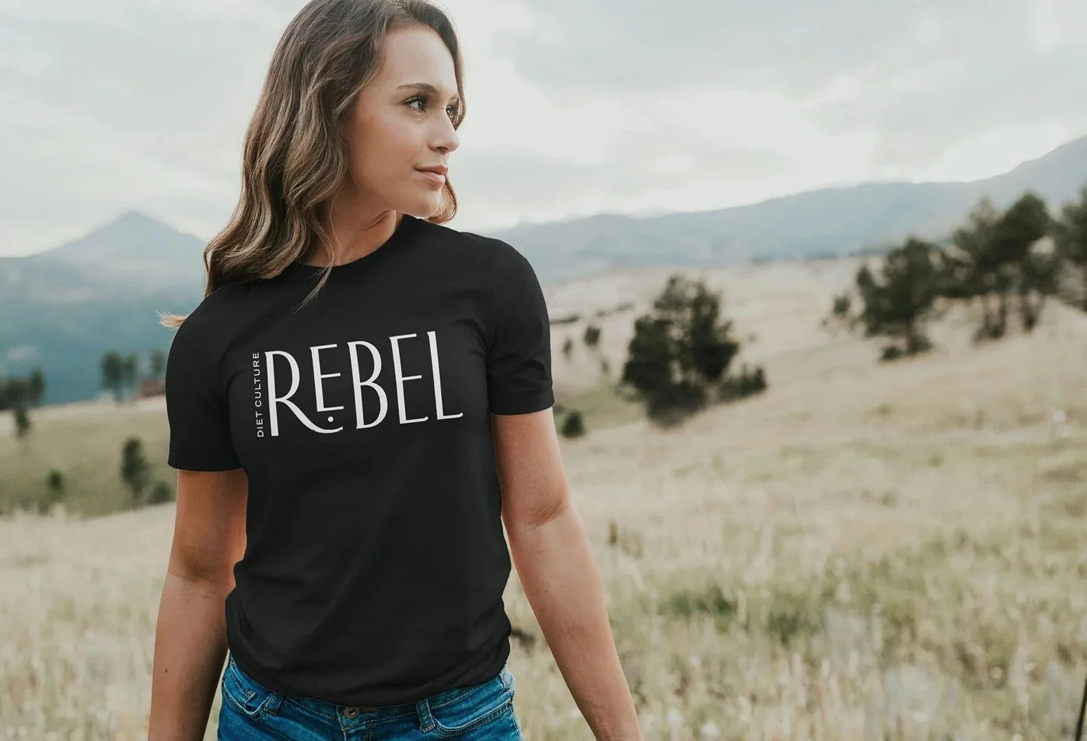 A woman wearing a black T-shirt with the words 'REBEL' and 'DIET CULTURE' written on it stands outdoors in a field with hills and trees in the background.