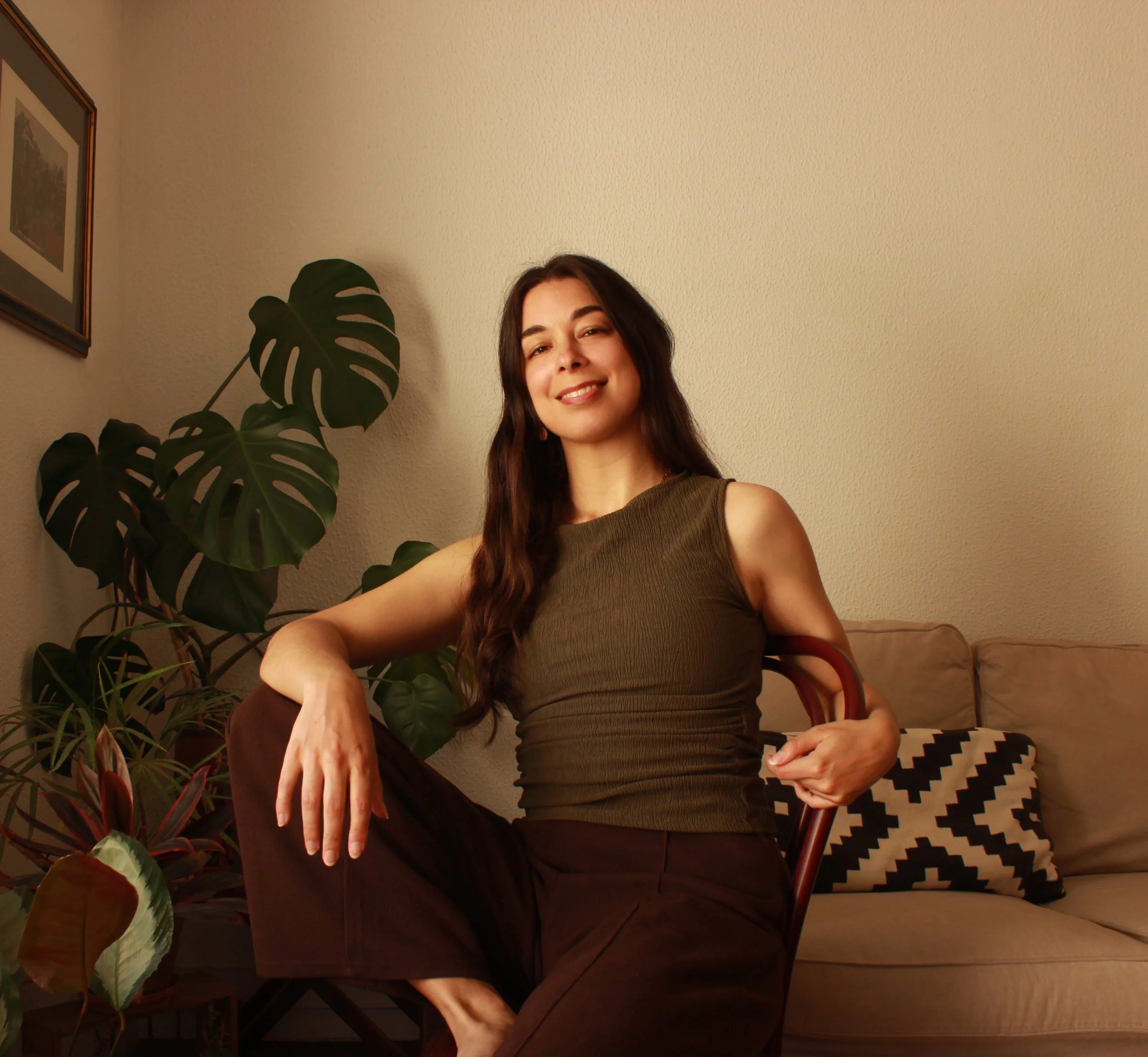 Inês Pessoa, a senior designer from Portugal and the founder of Wild Yolks - a boutique brand design studio. A woman with long, dark hair sitting in a chair, smiling at the camera, in a room with houseplants and framed pictures on the wall.