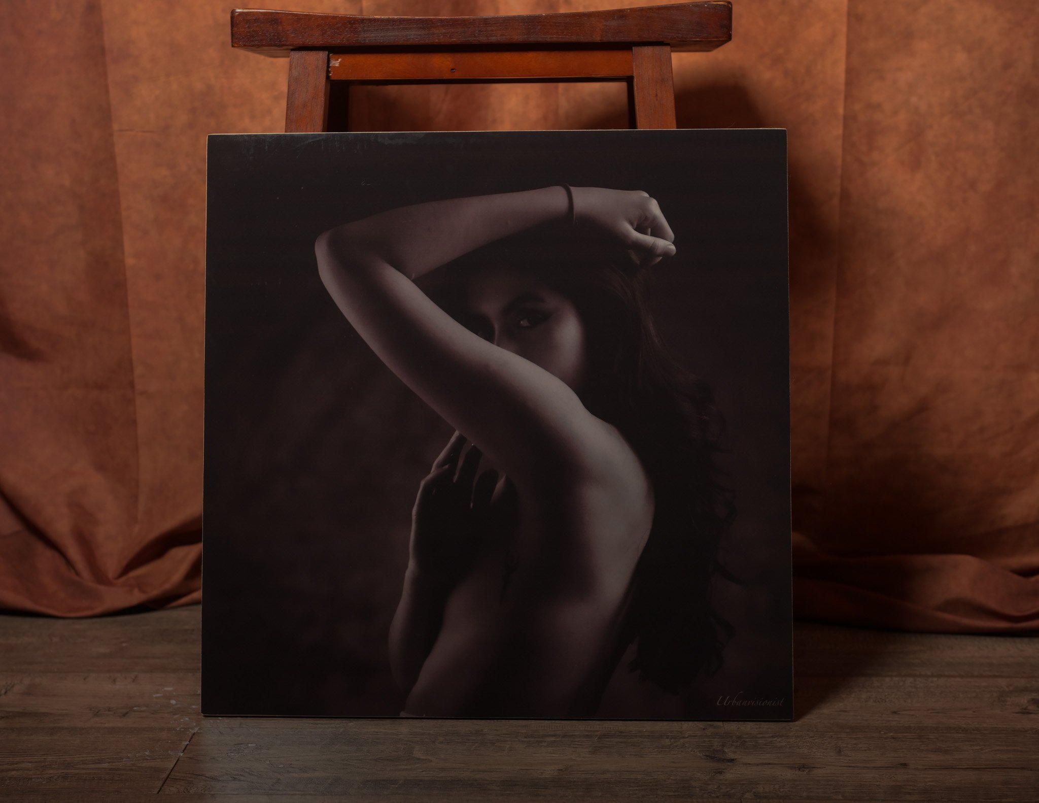 A black-and-white portrait photograph of a woman, emphasizing her arm and face partially obscured, set against a dark background and placed on a wooden surface with a wooden chair behind it.