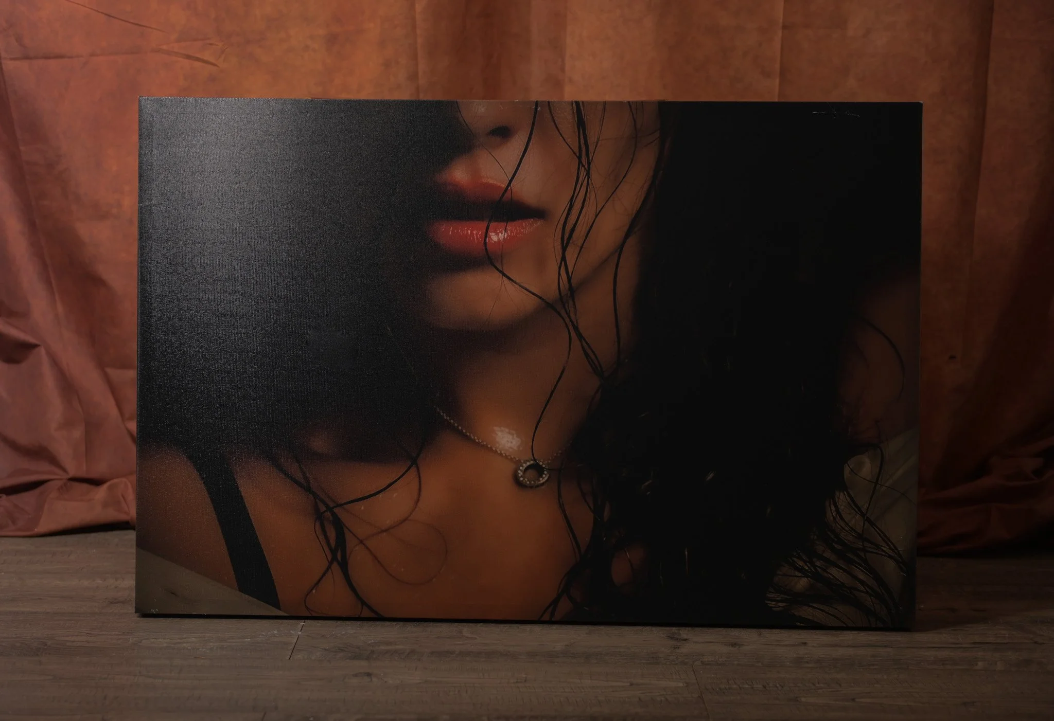 A close-up of a large canvas featuring a portrait of a woman with wet, dark hair, red lips, and a black strap visible on her shoulder. The woman is wearing a silver necklace with a circular pendant. The canvas is set against a wooden backdrop.