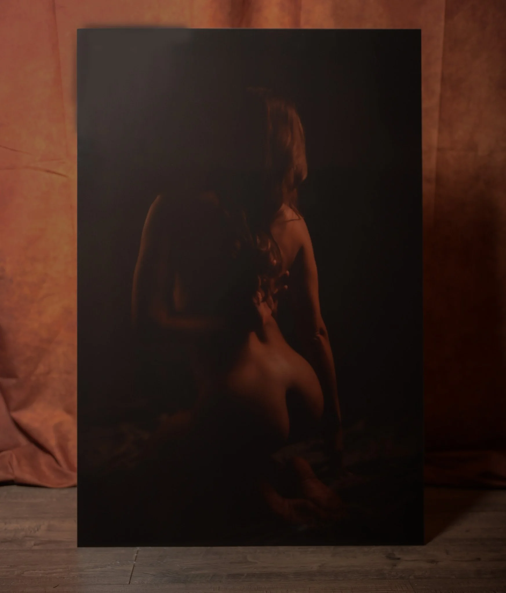 Dark, moody photograph of a nude woman with long hair, sitting with her back turned, illuminated by soft lighting.