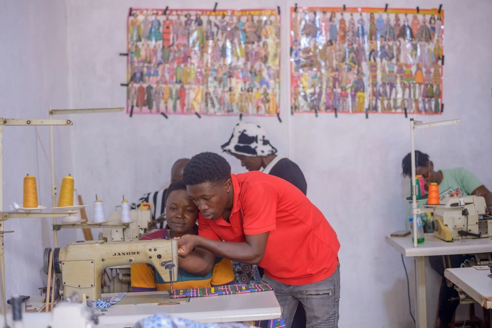 A sewing workshop with multiple workers using sewing machines; a man assists a woman at the foreground, and two women are working at sewing machines in the background, with colorful fabric and wall posters of various clothing designs.