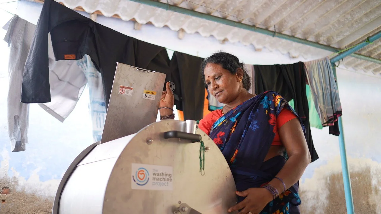 Woman in traditional Indian saree operating a washing machine outdoors with clothes hanging on a line behind her.