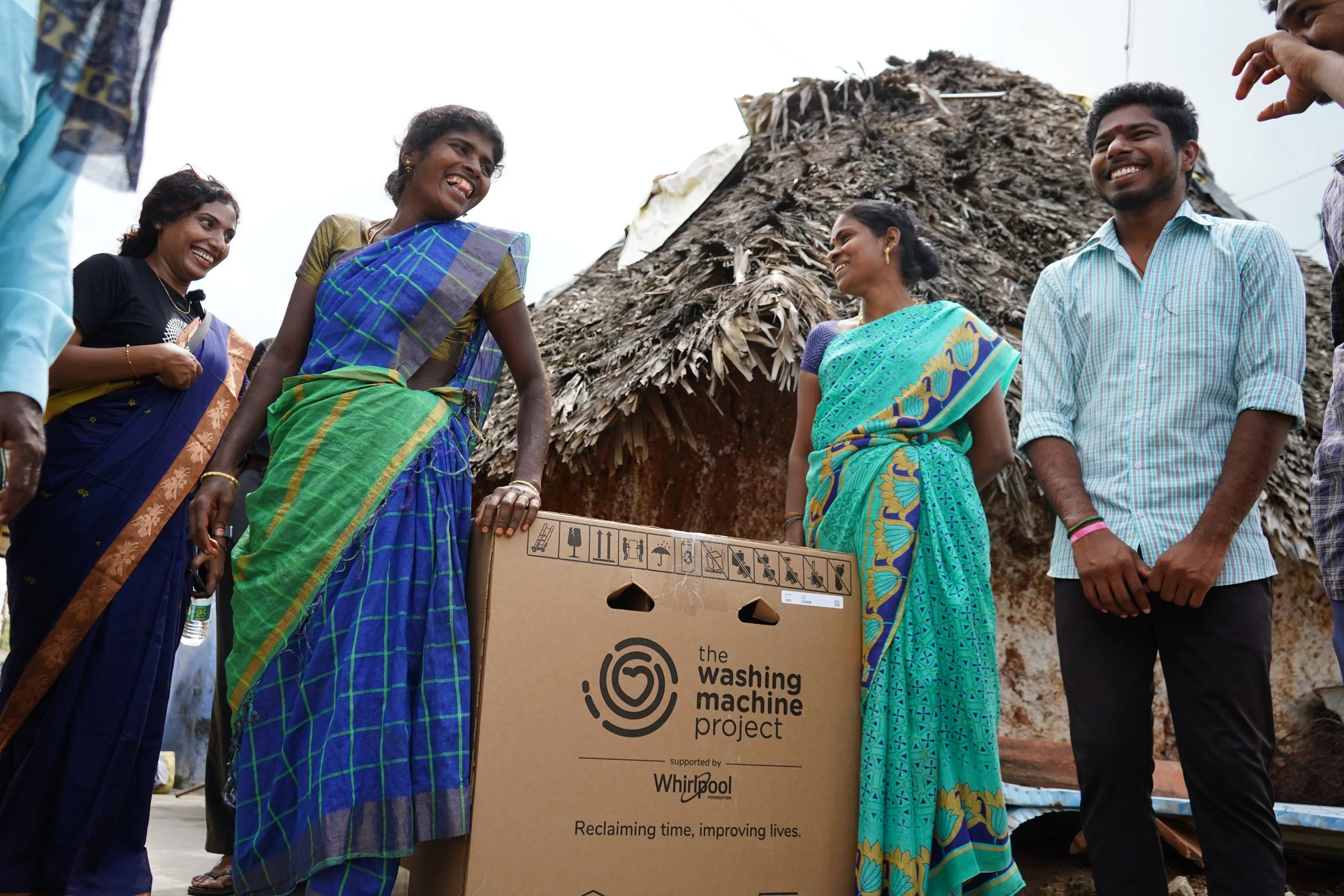 Group of people, mostly women, smiling and gathering around a cardboard box labeled "the washing machine project," supported by Whirlpool, in front of a thatched roof hut.