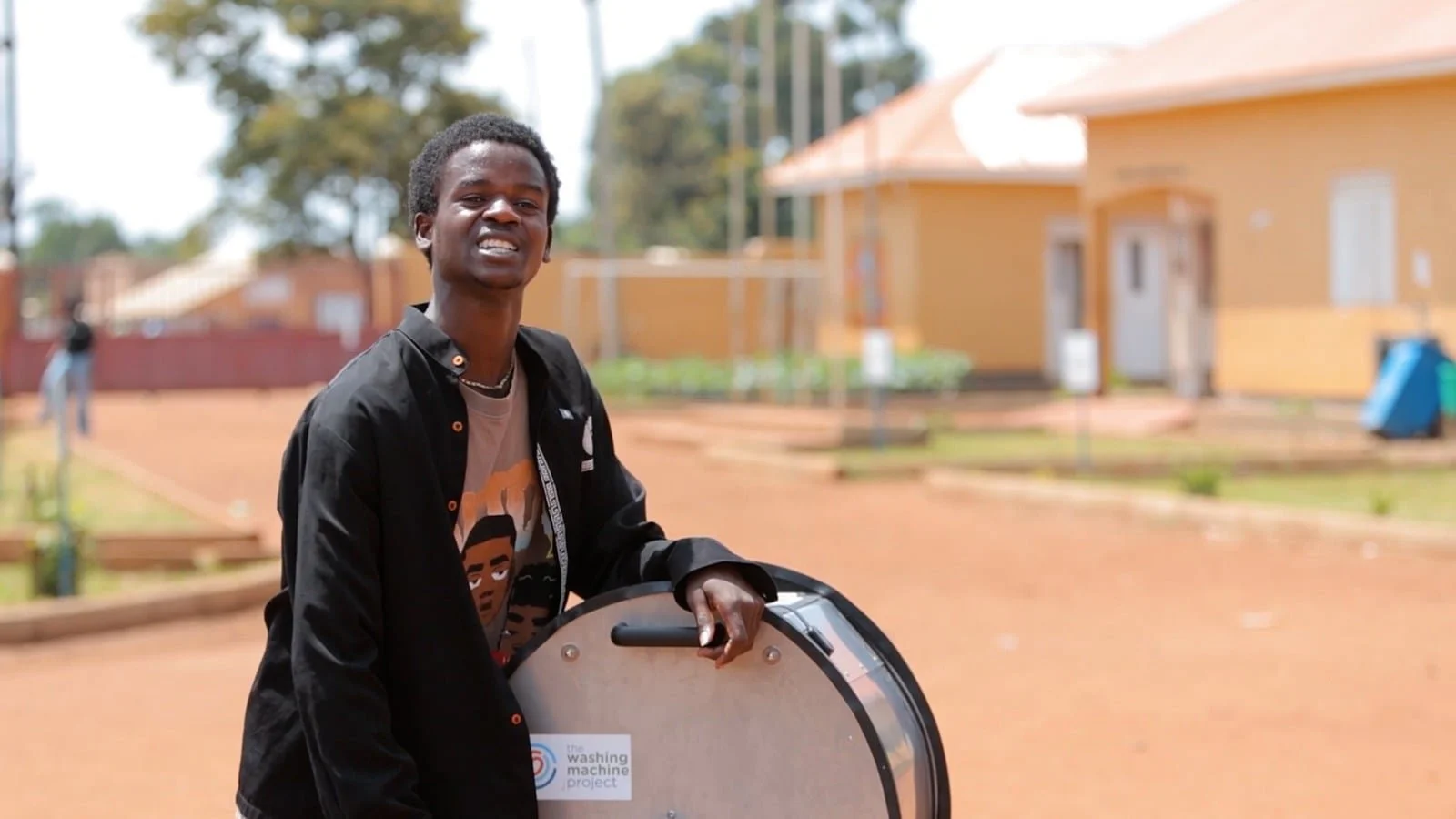 Young man with dark skin and short curly hair stands outside holding a round, metallic object with a label. He wears a black jacket over a graphic T-shirt and smiles. Background includes a dirt pathway, eco-friendly street furniture, and yellow buildings with white windows.