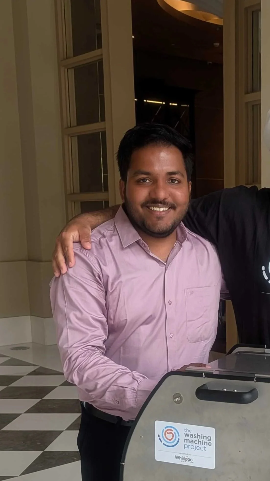 A smiling man in a pink shirt standing next to a washing machine promoting 'the washing machine project' supported by Whirlpool.