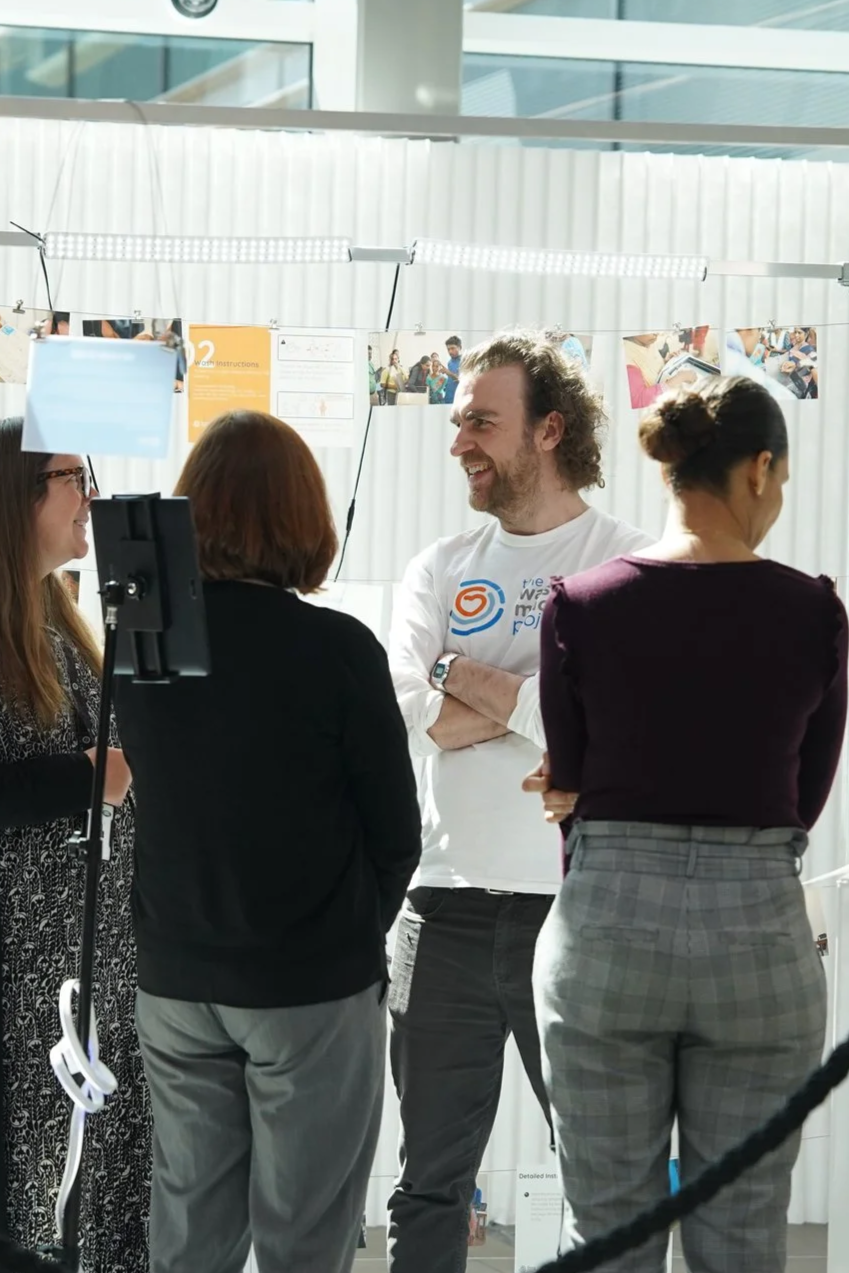 Four people are standing in a semi-circle, engaged in conversation at an indoor event. One man with curly hair and a beard is smiling, wearing a white long-sleeve shirt with a logo. The other three women have different hair styles and are dressed in casual and business attire. Behind them, there are posters and photographs hanging on a string, with a white, translucent background and large windows letting in natural light.