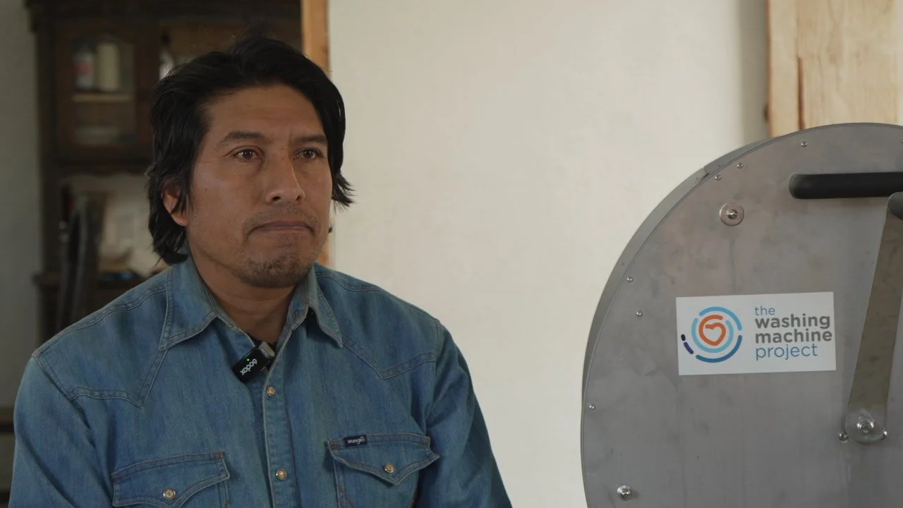A man with black hair and a serious expression standing near a washing machine with a "the washing machine project" sticker.