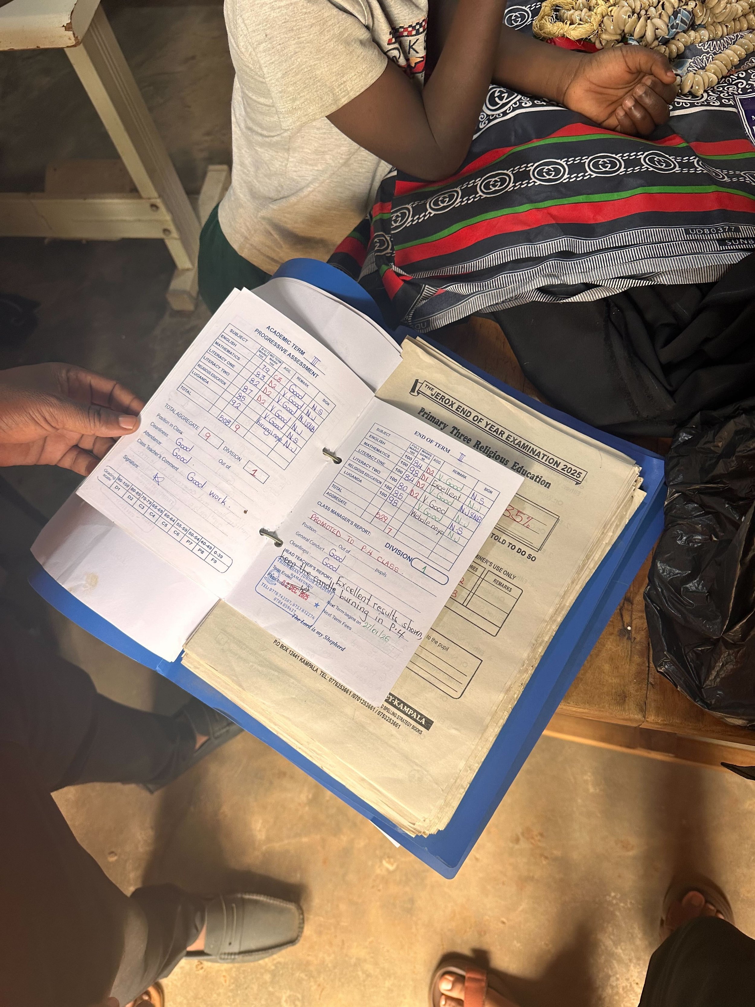 Photo of a person holding a report card with grades and comments, sitting at a desk with another person nearby, and a folder labeled 'End of Year Examination 2025' visible in the scene.