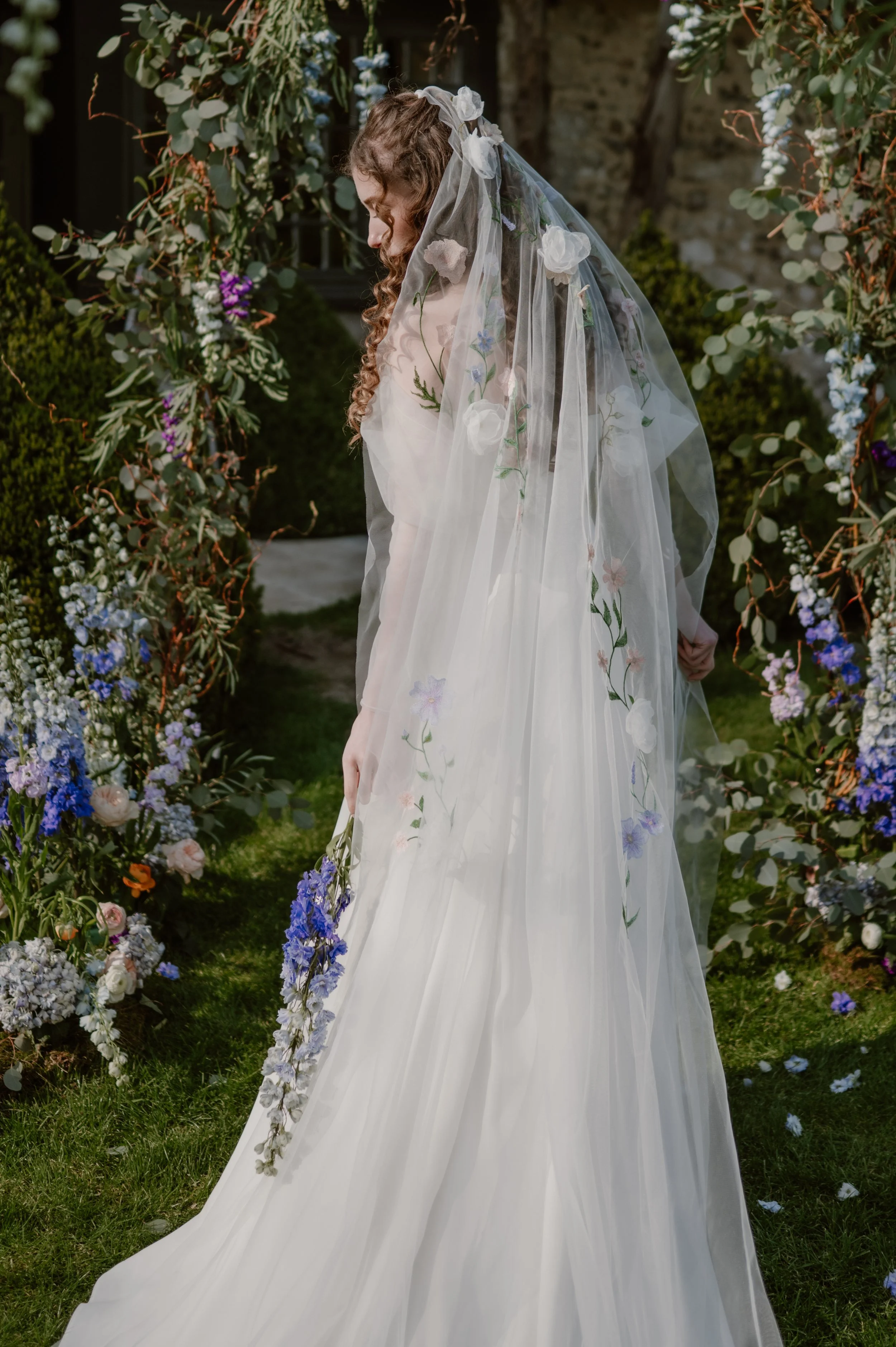 An image of a bride surrounded by an arch of blue and lilac flowers wearing a simple gown and a delicate wedding veil filled with embroidery and 3d flowers