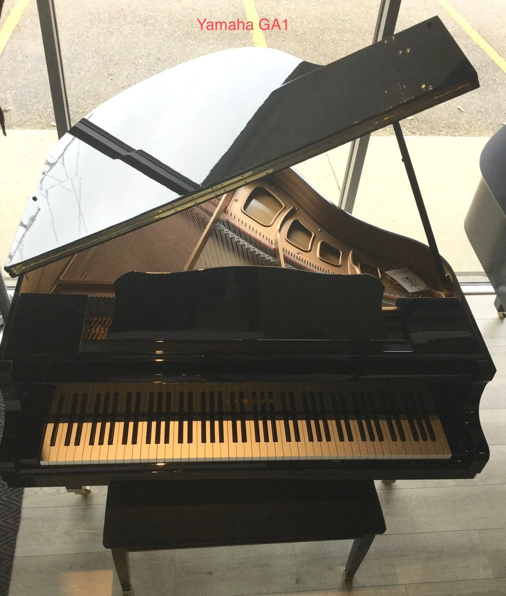 Black grand piano with open lid and matching bench in a store with large glass windows showing outdoor trees and parked cars.