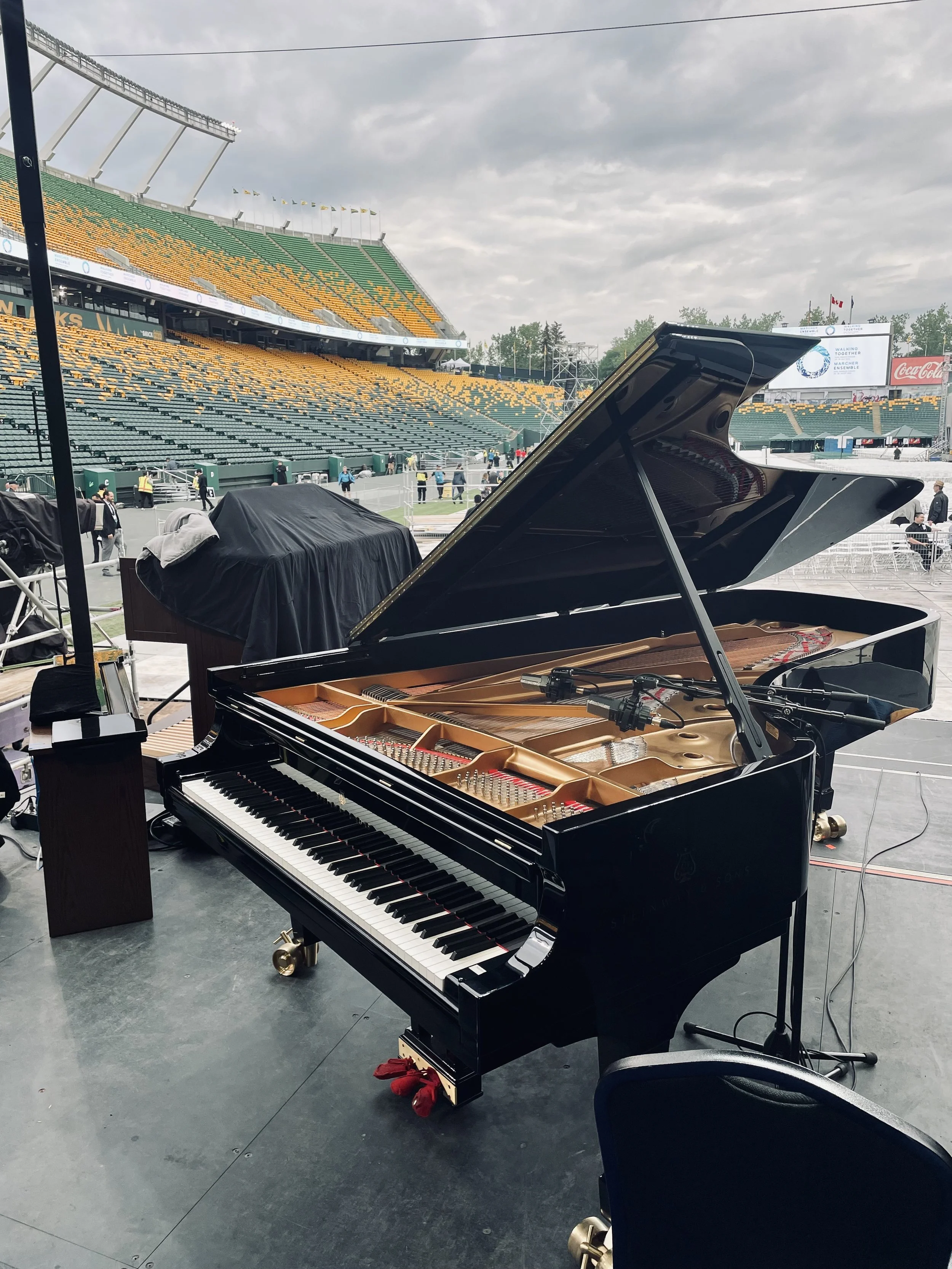 A black grand piano with its lid open on a stage at a stadium, prepared for a performance. The stadium seats are green and yellow, and the sky is cloudy.