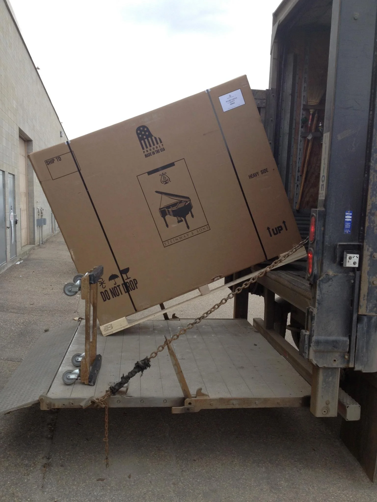 A large brown box loaded onto a flat truck bed, with a dolly and a chain securing it, outside a building.