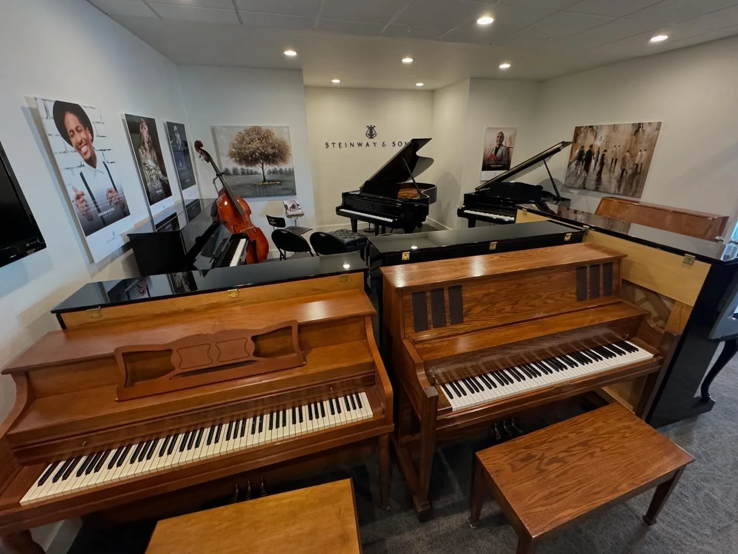 Multiple pianos of different styles and wood finishes arranged in a music store or showroom, with framed posters and paintings on the wall.