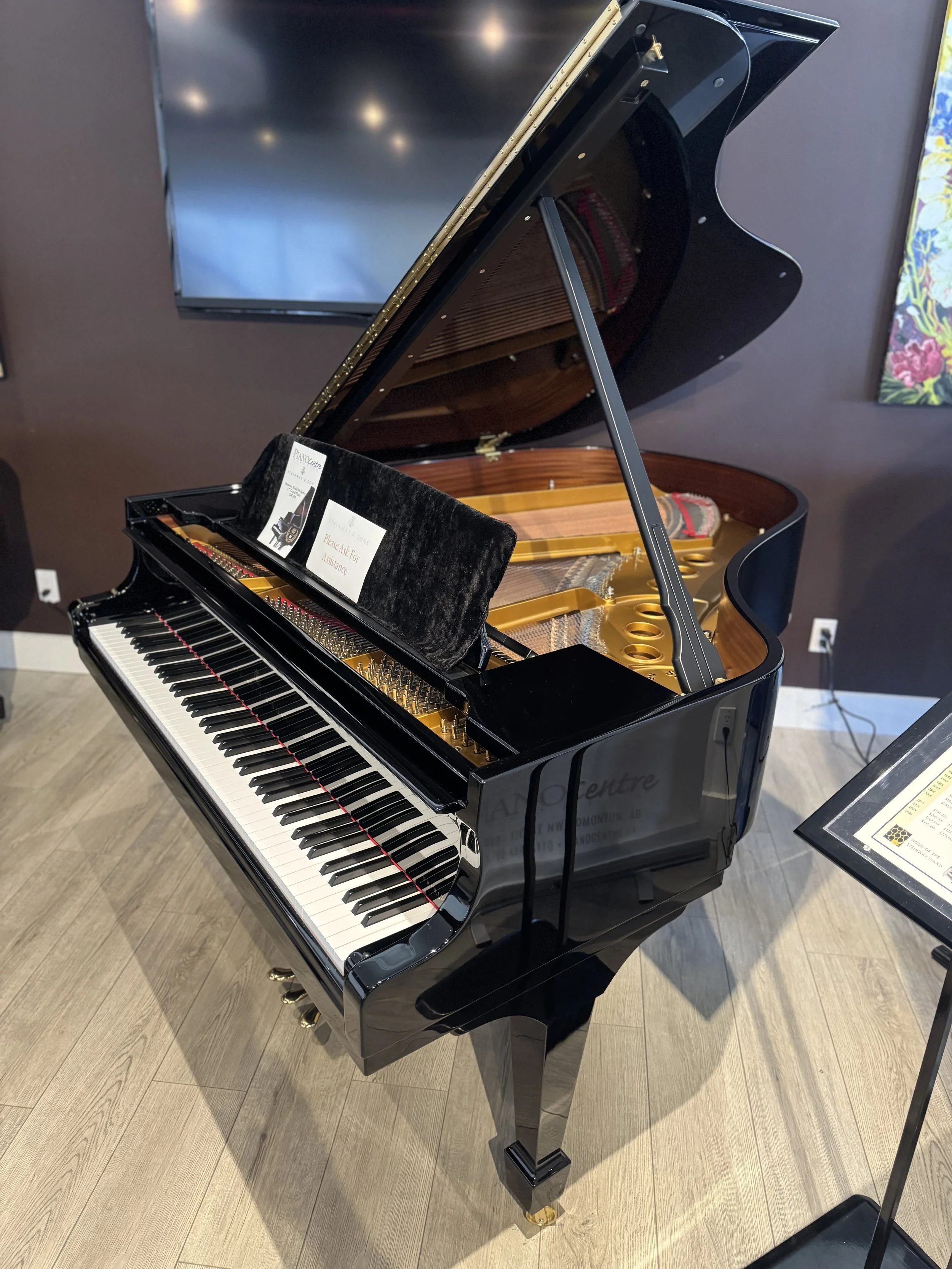 A Steinway & Sons grand piano with an open lid, showing the interior strings and hammers, situated in a room with a wooden floor and a white wall.